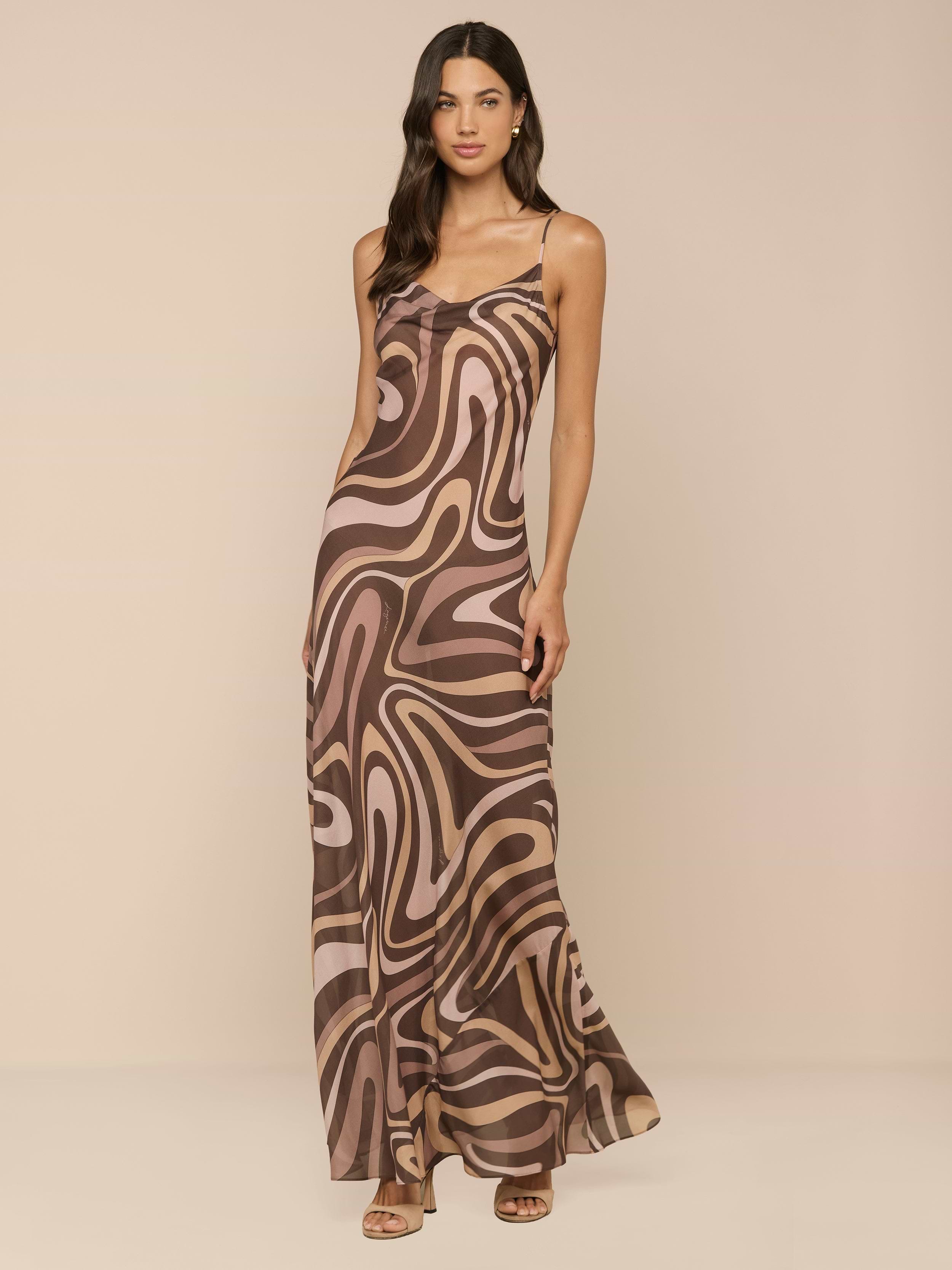 A woman wearing the Christine Silk Gown, a brown swirl print sleeveless maxi dress with spaghetti straps, styled with sandals and standing against a plain white background.