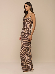 A woman stands sideways against a plain white background, wearing the Christine Silk Gown—a sleeveless maxi dress in a brown swirl print.