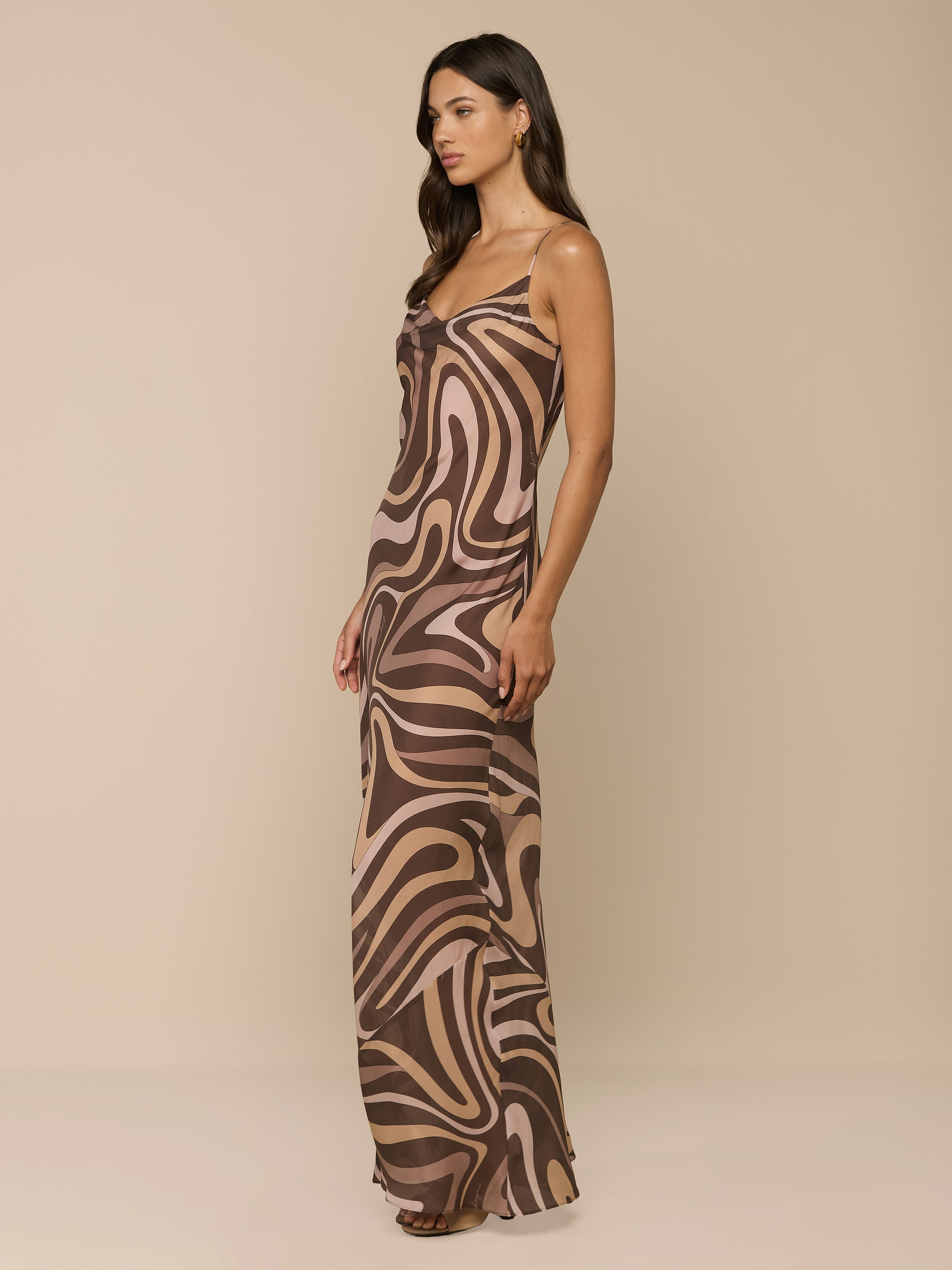 A woman stands sideways against a plain white background, wearing the Christine Silk Gown—a sleeveless maxi dress in a brown swirl print.