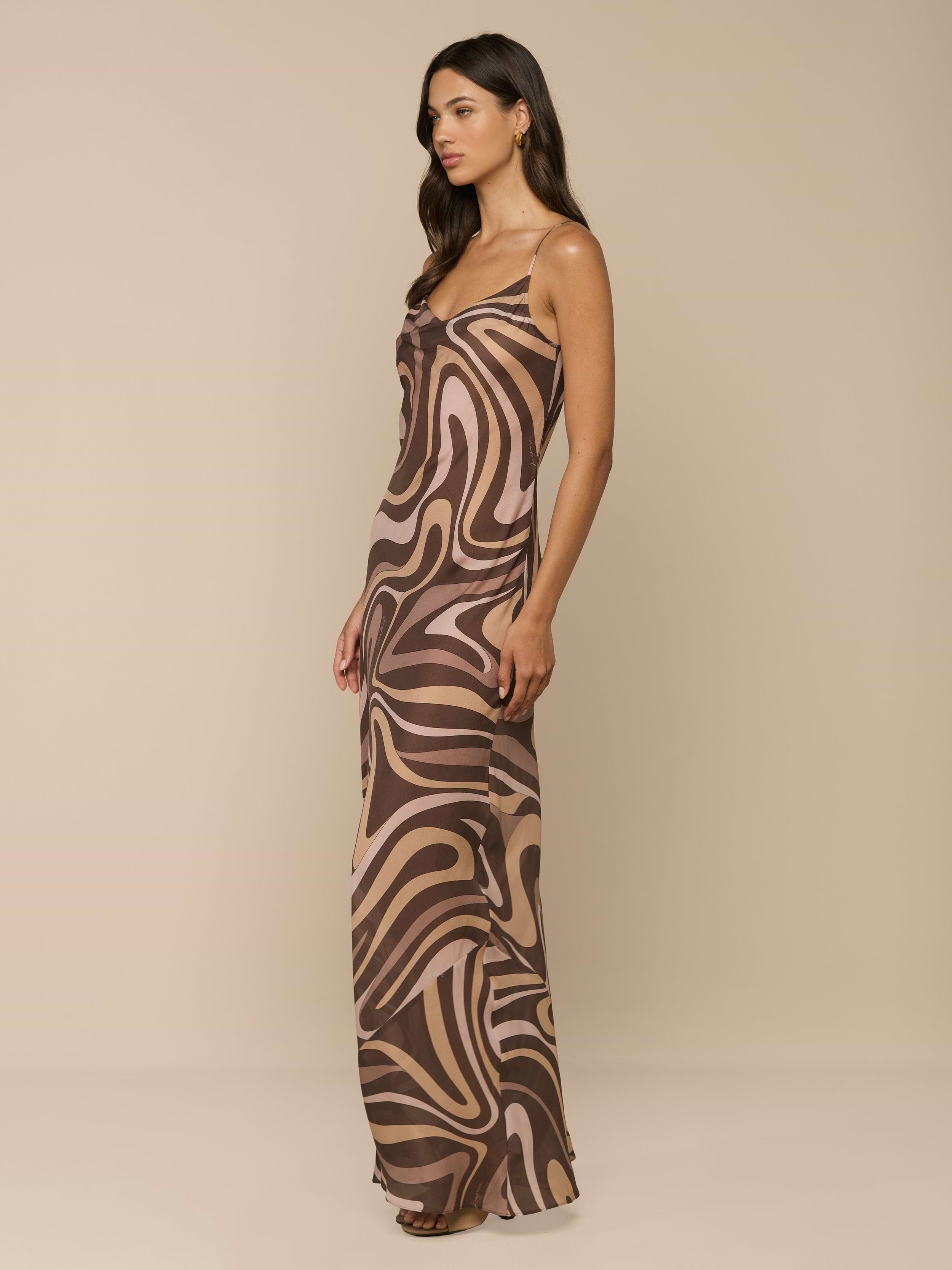 A woman stands sideways against a plain white background, wearing the Christine Silk Gown—a sleeveless maxi dress in a brown swirl print.