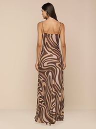 A woman stands facing away, wearing the Christine Silk Gown—a long, bias-cut dress with thin straps and a brown swirl print in shades of beige and pink—against a plain white background.