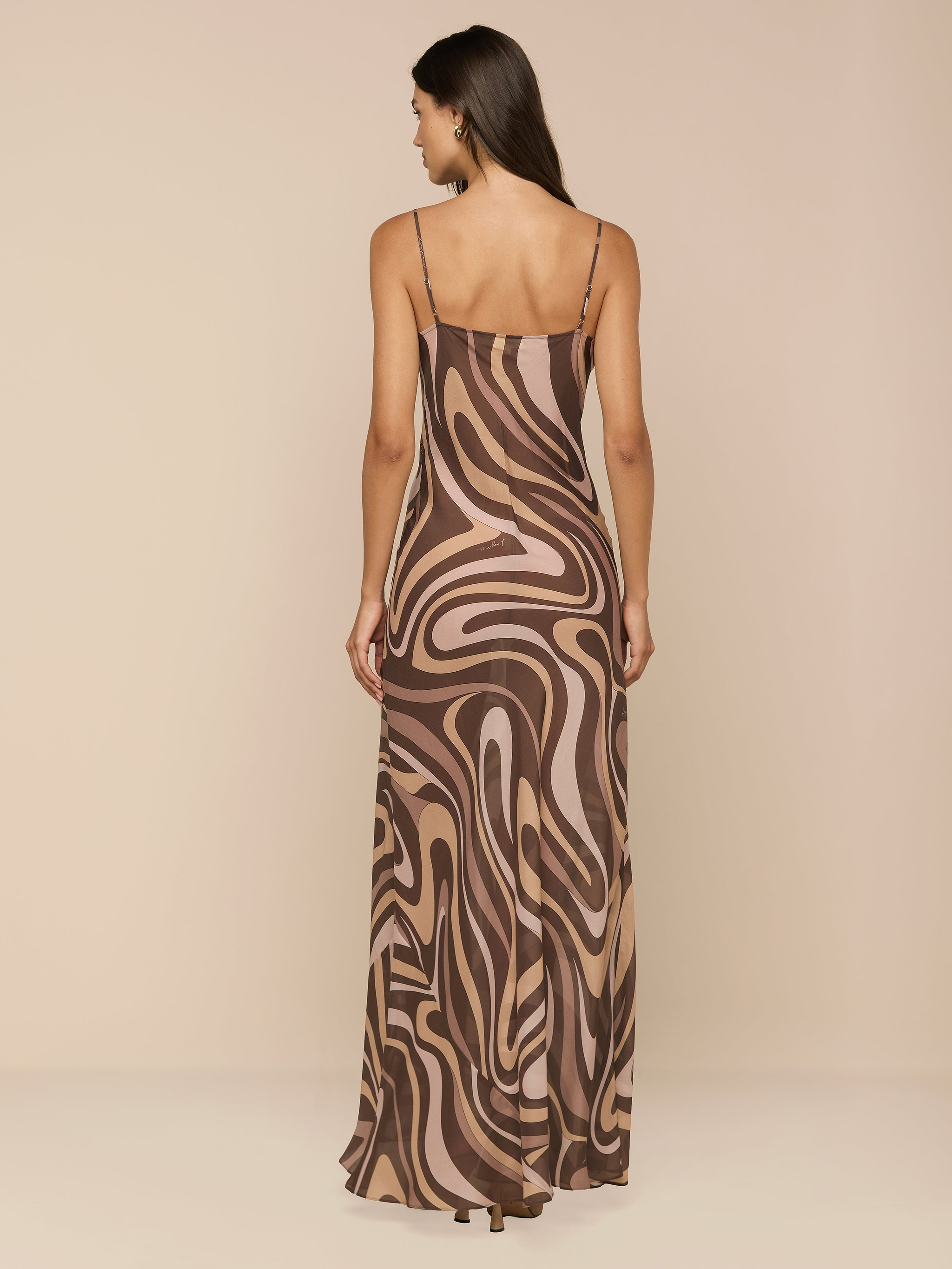 A woman stands facing away, wearing the Christine Silk Gown—a long, bias-cut dress with thin straps and a brown swirl print in shades of beige and pink—against a plain white background.