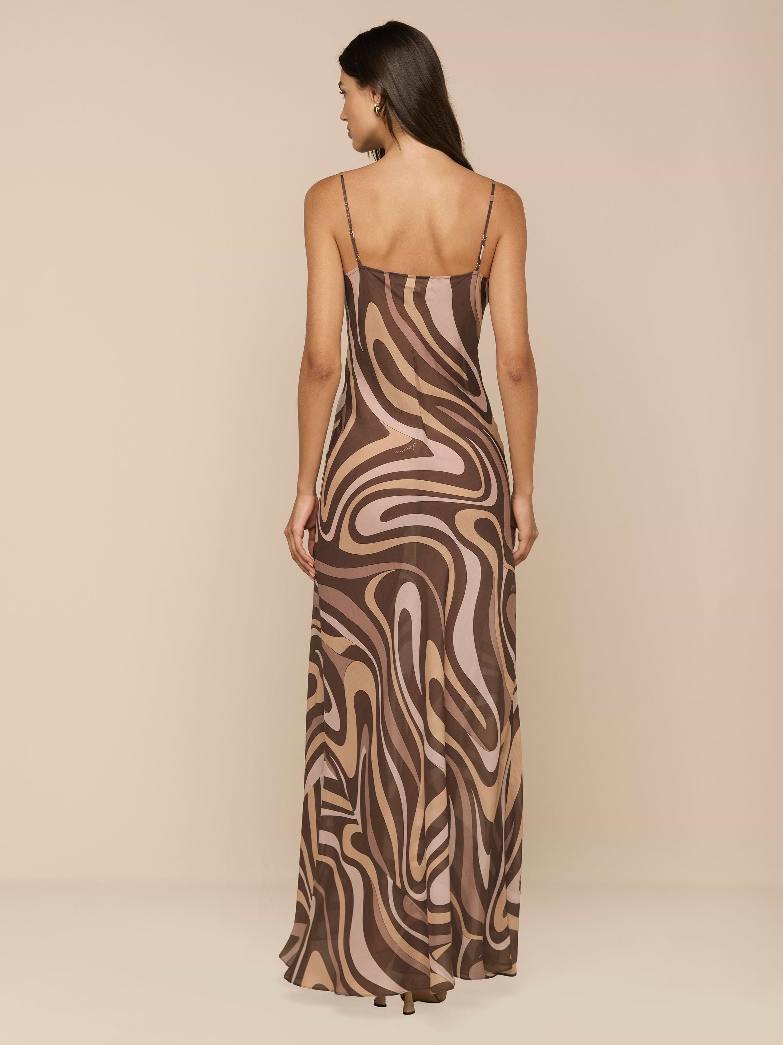 A woman stands facing away, wearing the Christine Silk Gown—a long, bias-cut dress with thin straps and a brown swirl print in shades of beige and pink—against a plain white background.