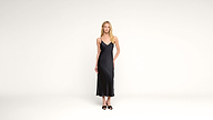 A woman stands in a minimalist white space wearing a black sleeveless midi dress and black sandals, looking straight at the camera with her arms relaxed at her sides.