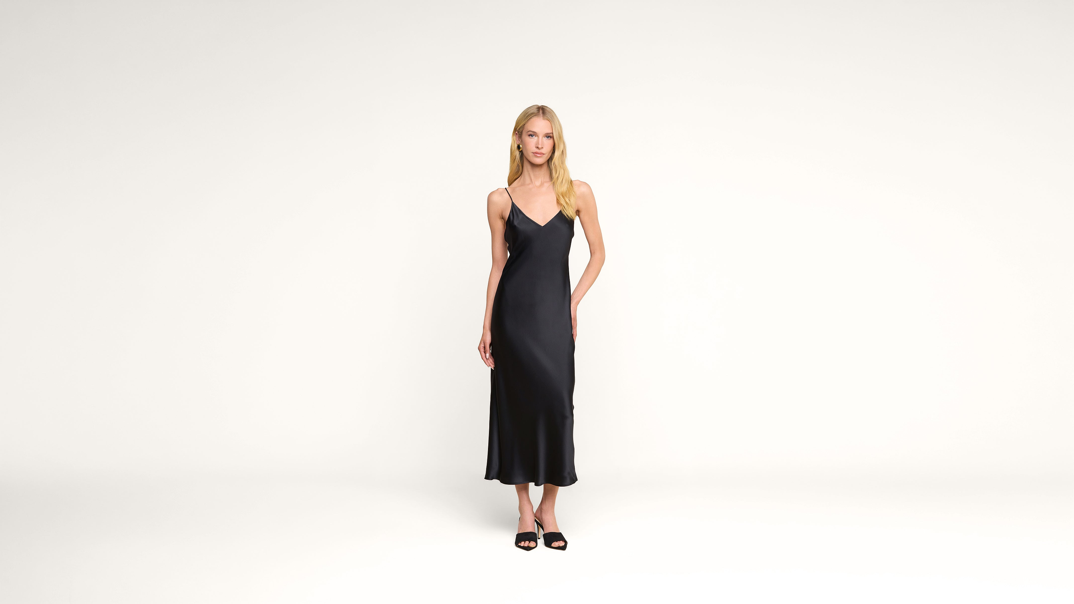 A woman stands in a minimalist white space wearing a black sleeveless midi dress and black sandals, looking straight at the camera with her arms relaxed at her sides.