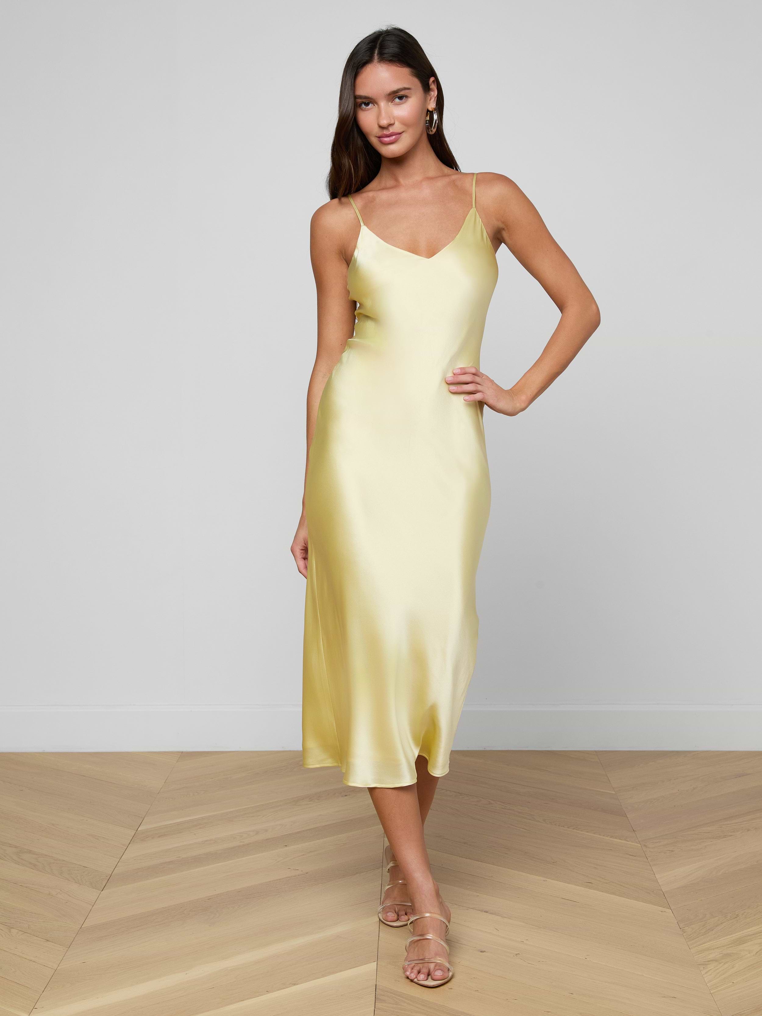 Wearing the Seridie Silk Slip Dress in yellow and beige sandals, a woman stands posing against a plain white background.