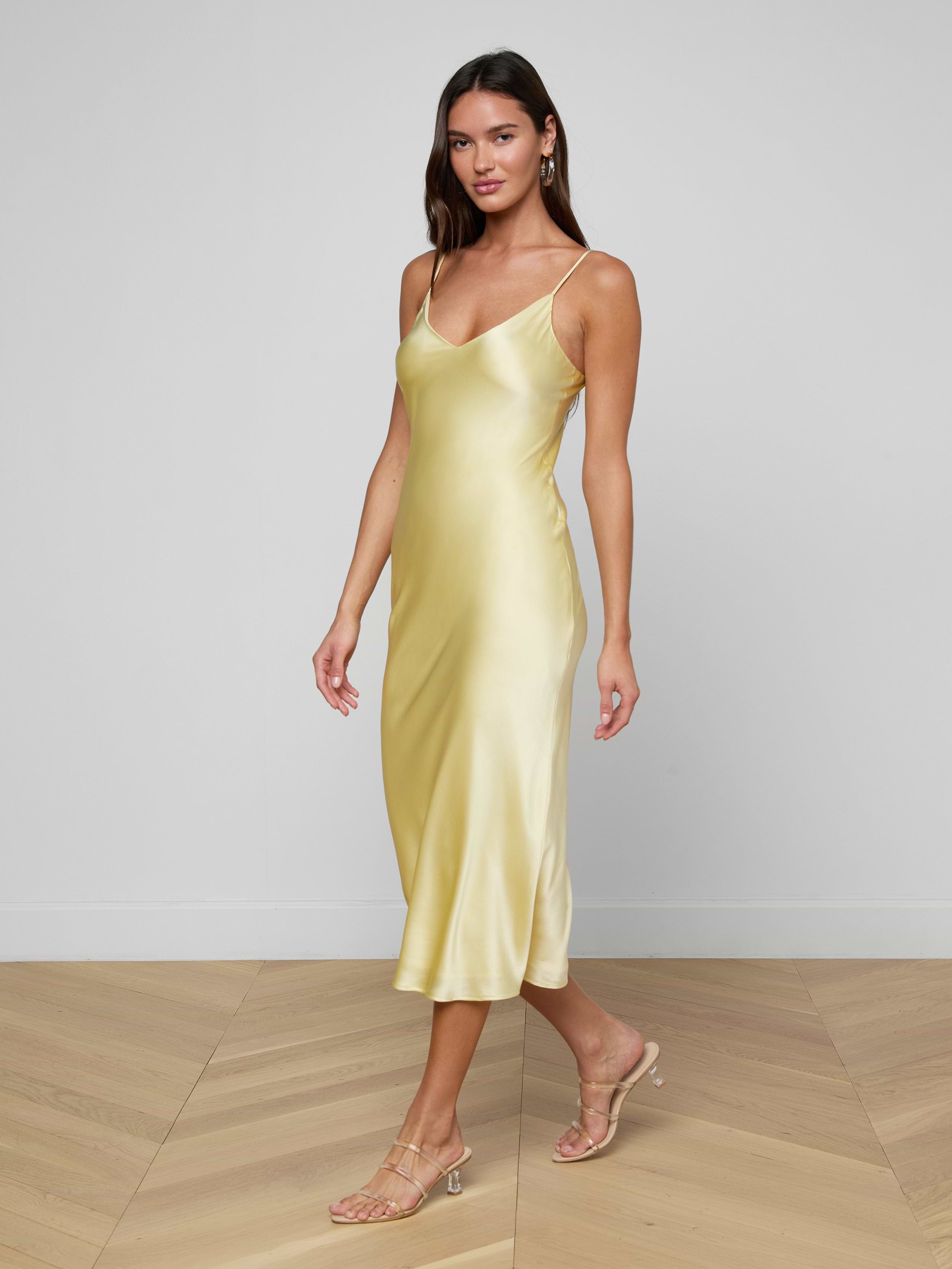 A woman wearing the Seridie Silk Slip Dress in yellow, featuring a flattering bias cut, paired with beige heeled sandals, walking against a plain white background.