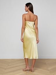 A woman with long hair stands facing away on a plain white background, wearing the Seridie Silk Slip Dress—a light yellow, bias-cut silk design with thin straps—paired with beige heeled sandals.