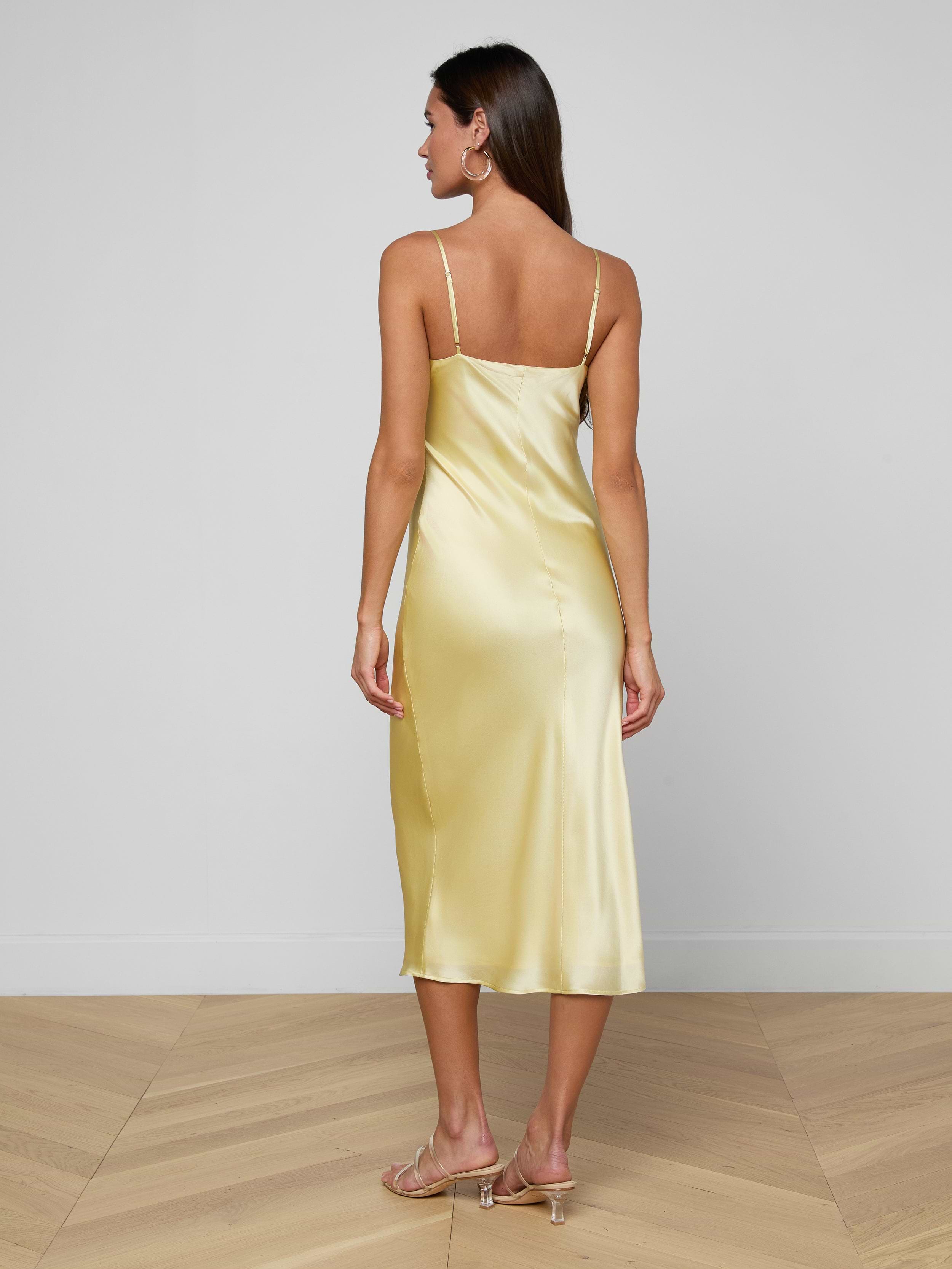 A woman with long hair stands facing away on a plain white background, wearing the Seridie Silk Slip Dress—a light yellow, bias-cut silk design with thin straps—paired with beige heeled sandals.