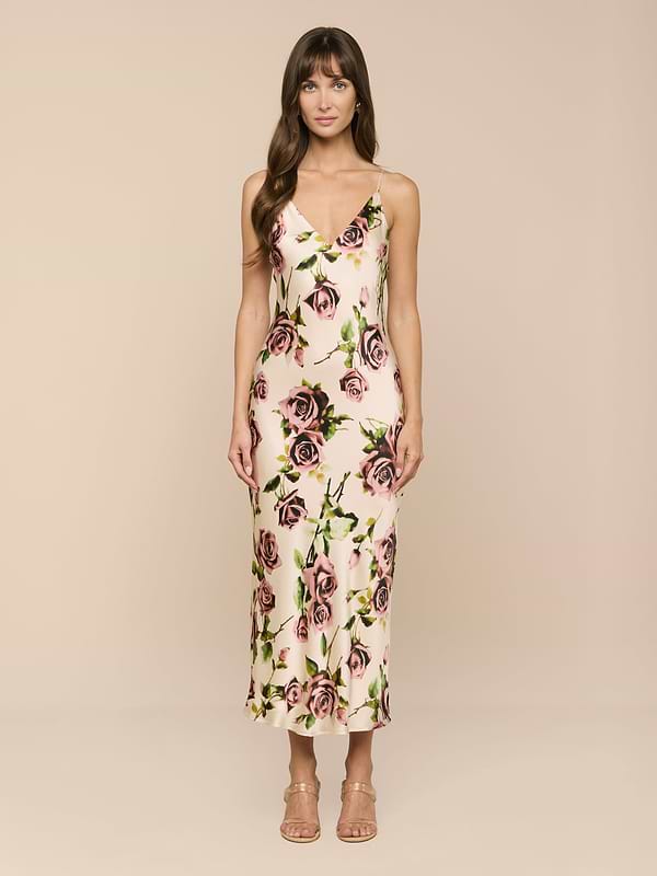A woman stands facing forward, wearing the Seridie Silk Slip Dress—a bias-cut, sleeveless V-neck midi dress featuring a pink and purple rose print. She has long brown hair and is wearing heeled sandals against a plain white background.