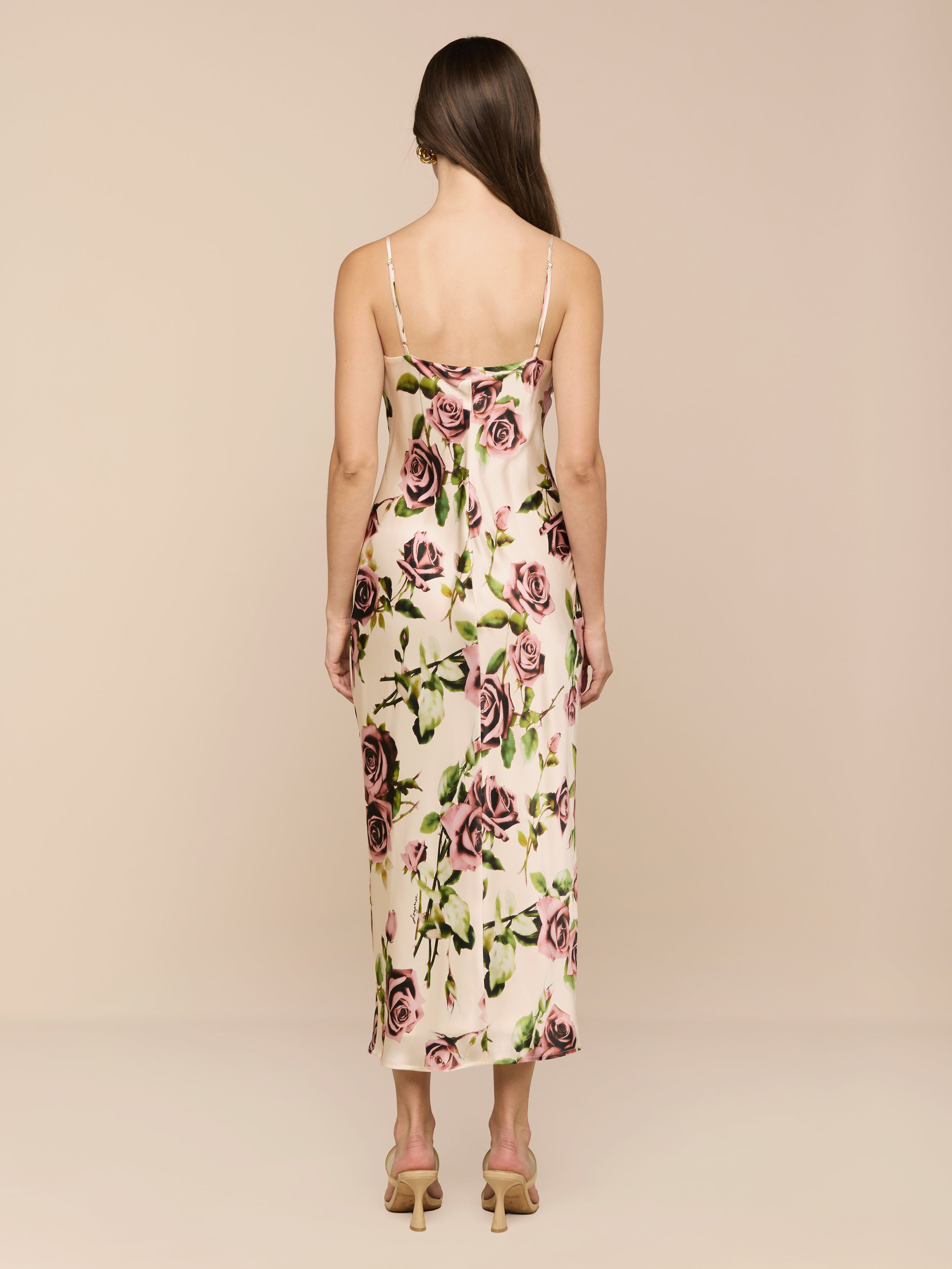 A woman with long brown hair stands against a plain white background, shown from the back, wearing the Seridie Silk Slip Dress—a bias-cut silk slip dress with a rose print—paired with heeled sandals.