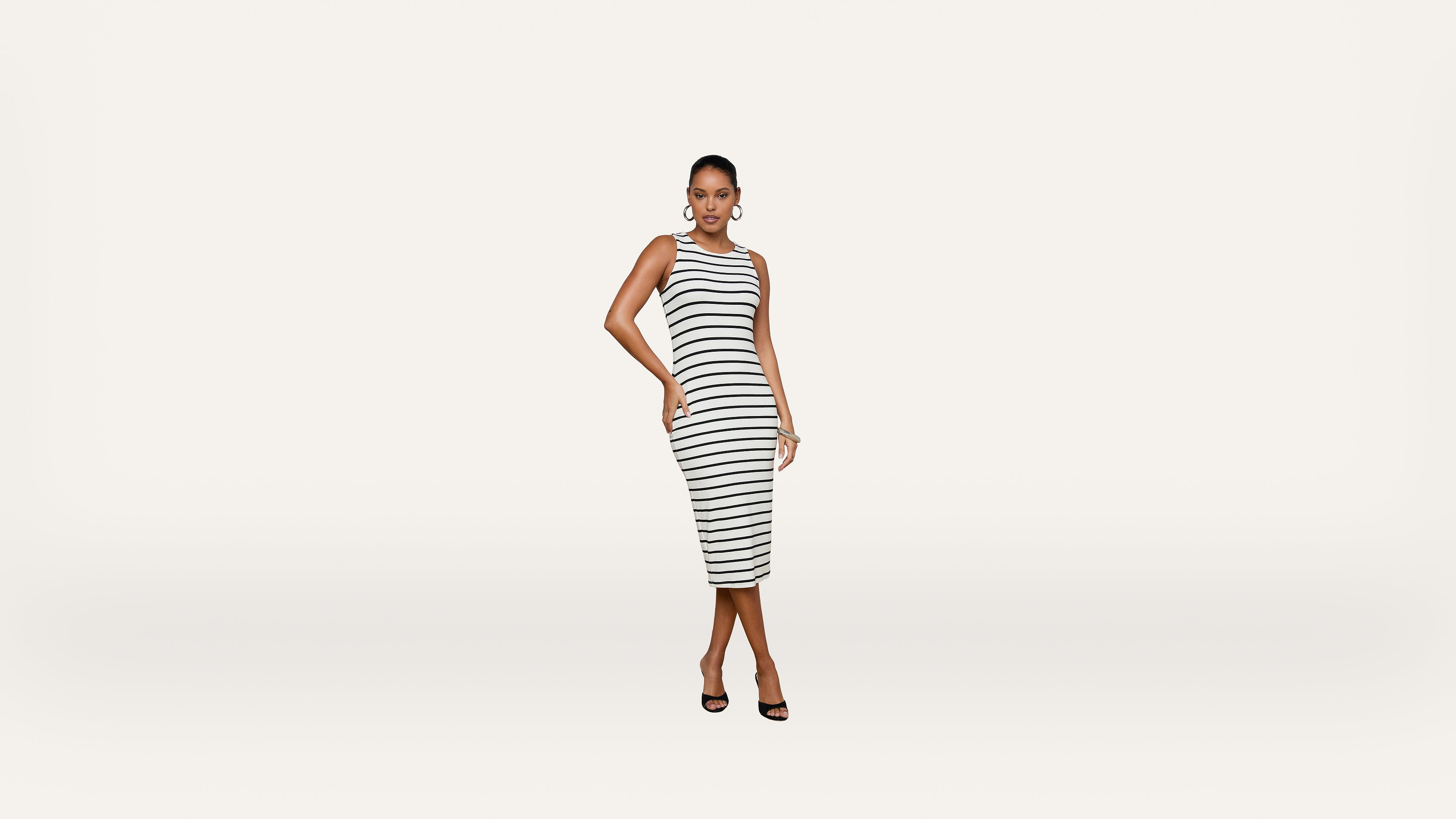 Woman wearing a sleeveless, knee-length, black and white striped dress and black heels stands against a plain white background, posing with one hand on her hip.