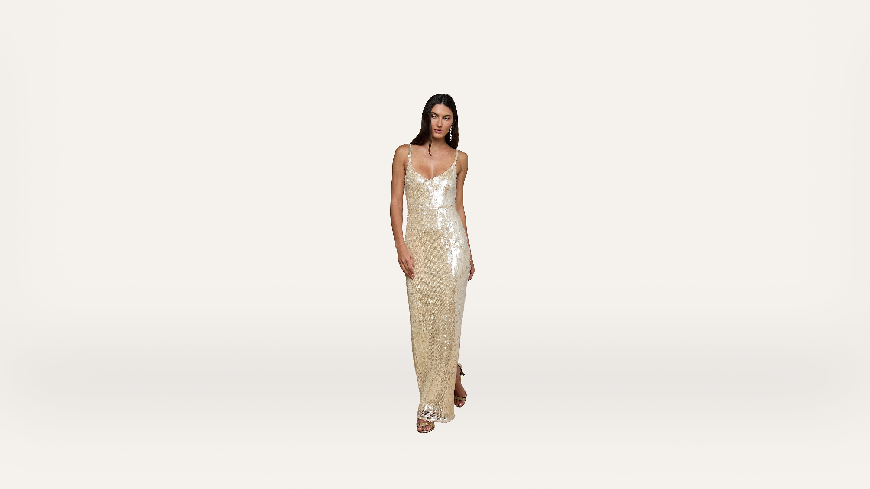 A woman wearing a sleeveless, floor-length, sparkling gold dress with thin straps stands against a plain, light background.