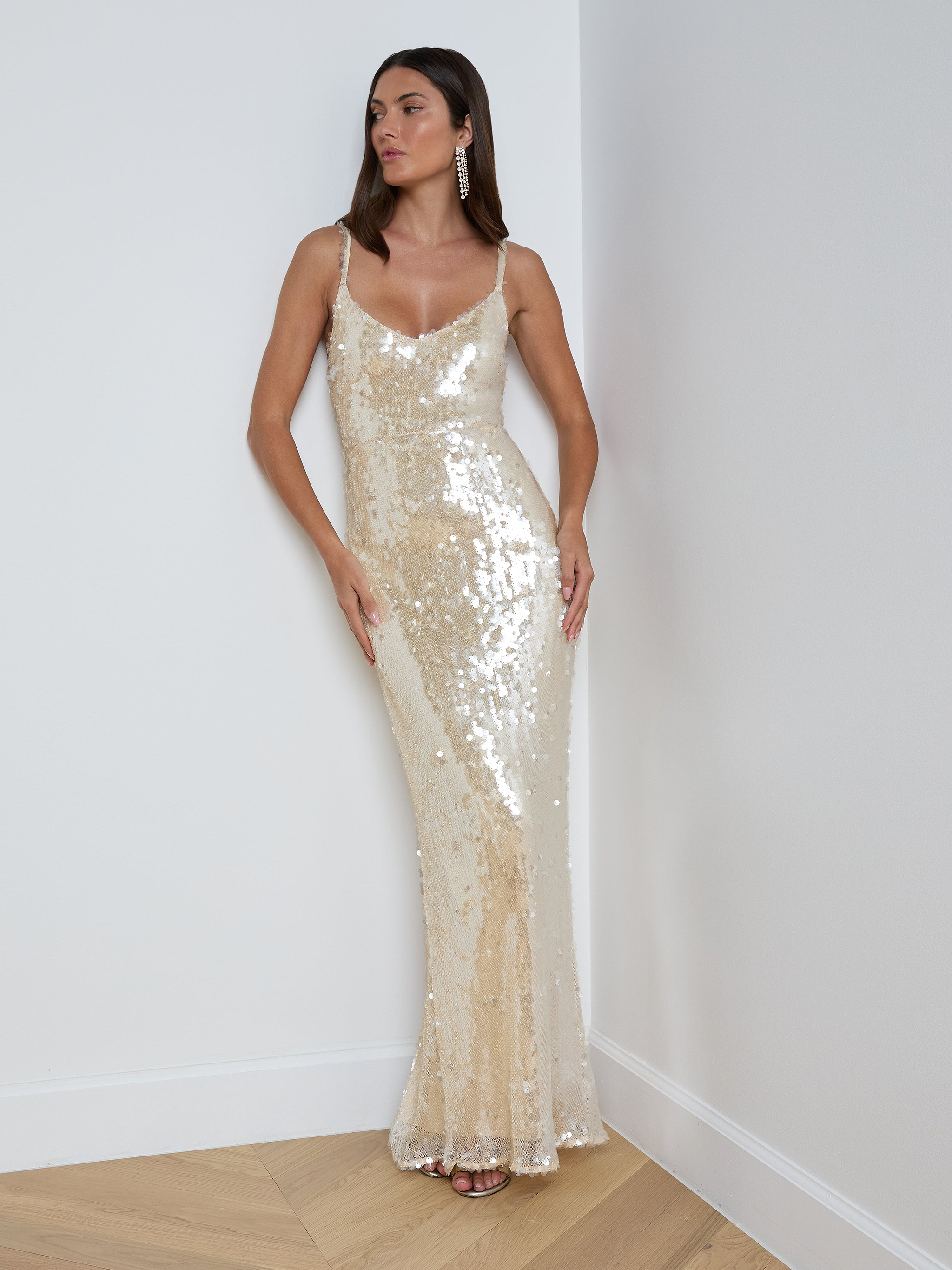 A woman wearing the Karma Sequin Maxi Dress—a sleeveless, floor-length gold dress featuring shimmering sequins and a V-neckline—stands against a plain white background. This elegant gown is perfect for NYFW 2026.