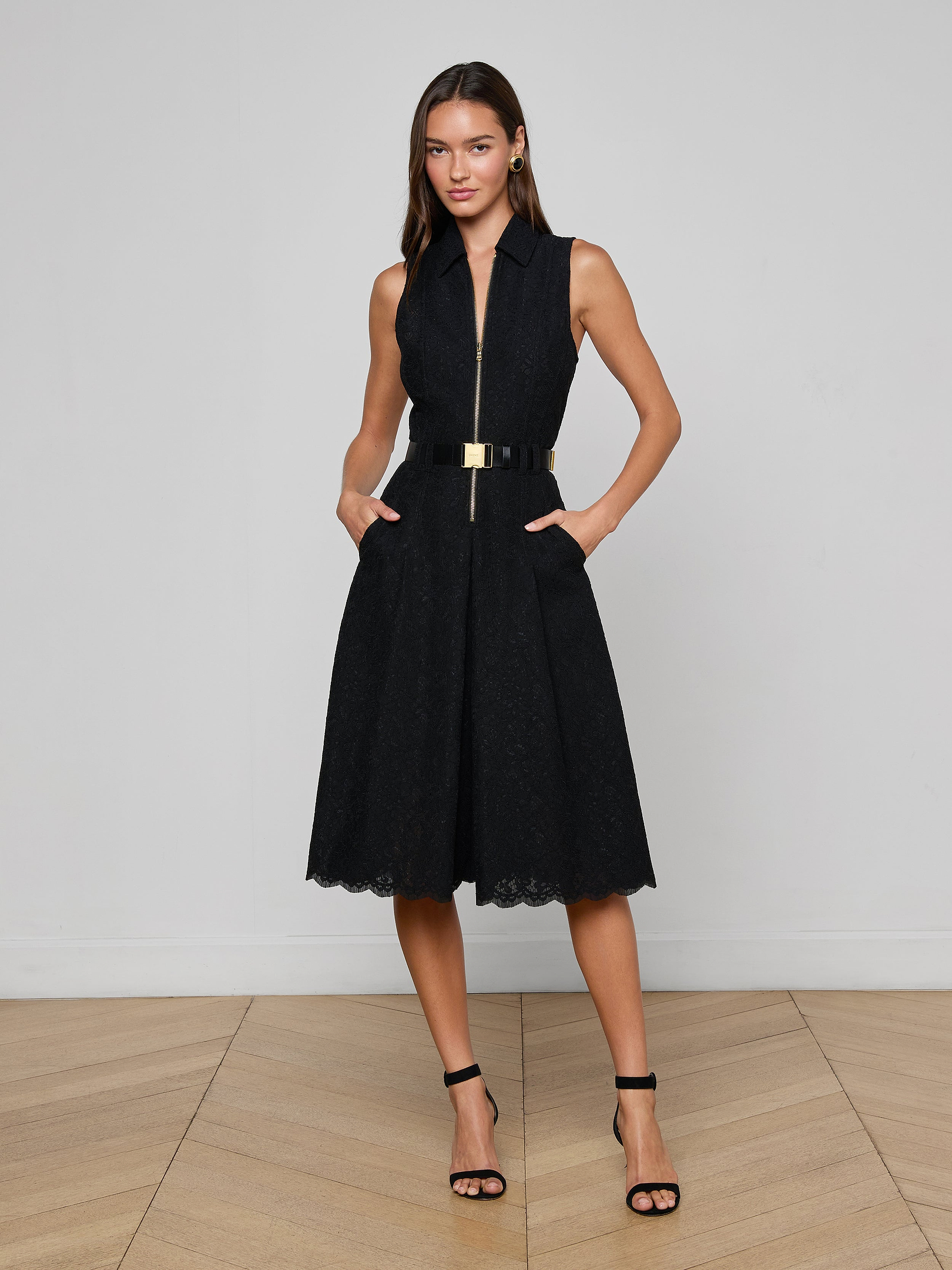 A woman stands against a plain white background wearing the Kylo Lace Shirt Dress—a sleeveless black knee-length dress with a collared neckline, gold belt, and black high-heeled sandals—perfect for an elegant desk-to-drinks look.