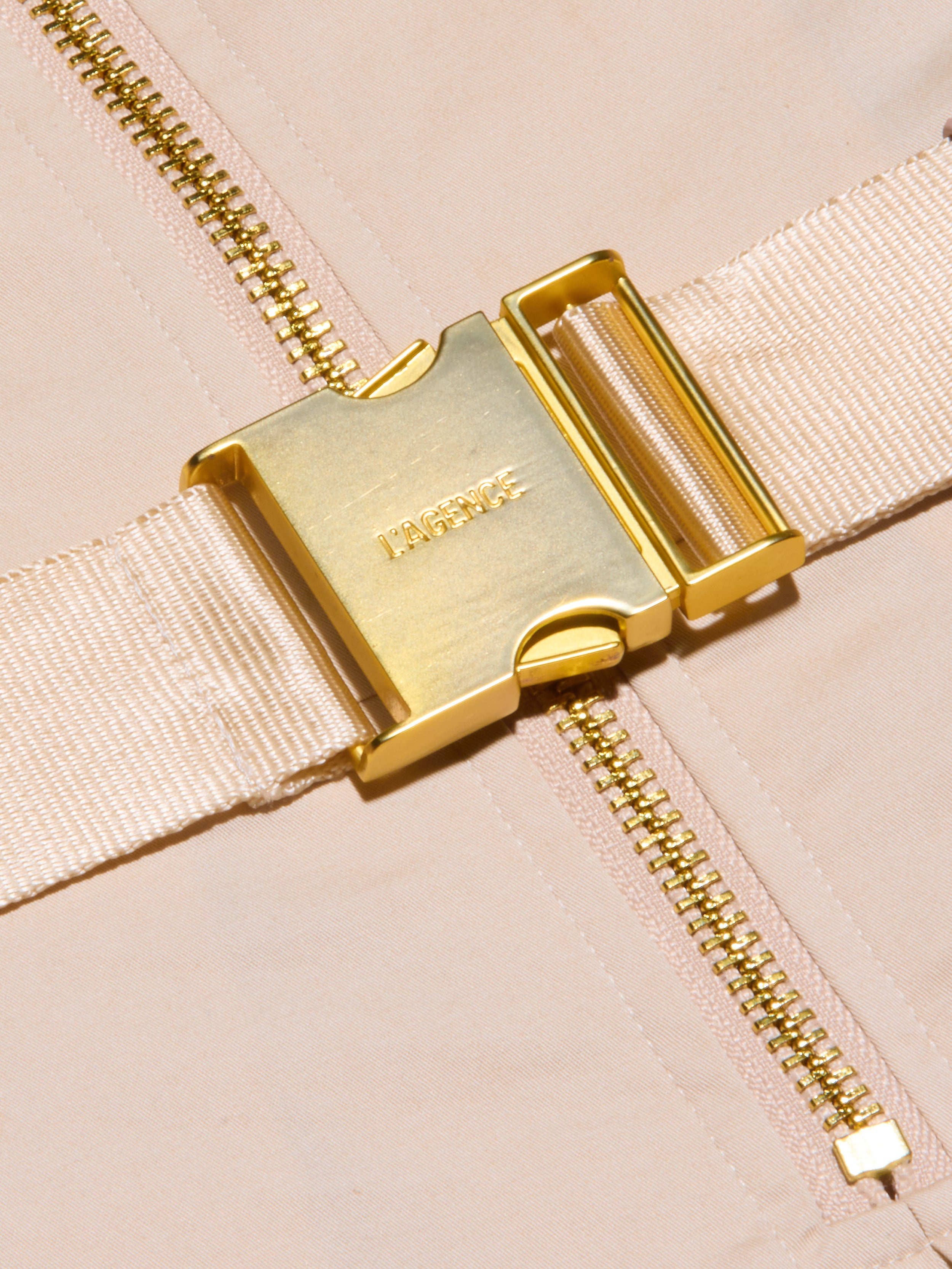 Close-up of a shiny gold buckle engraved with LAGENCE, attached to a beige fabric strap—part of the Kylo Poplin Shirt Dress—lying across a gold zipper on a light beige background.