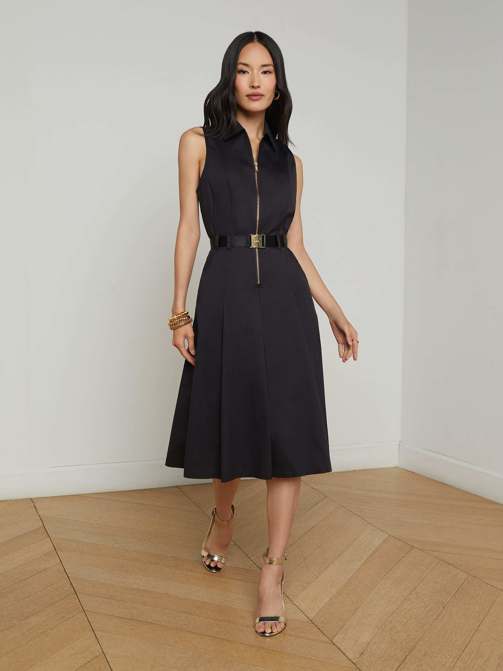 A woman in the Kylo Poplin Shirt Dress—a sleeveless black dress with a belt—stands against a plain white background, styled with gold heels and bracelets. Her hair is down, and her hands are relaxed at her sides. This versatile piece offers effortless style for any occasion, seamlessly taking you from desk to drinks.