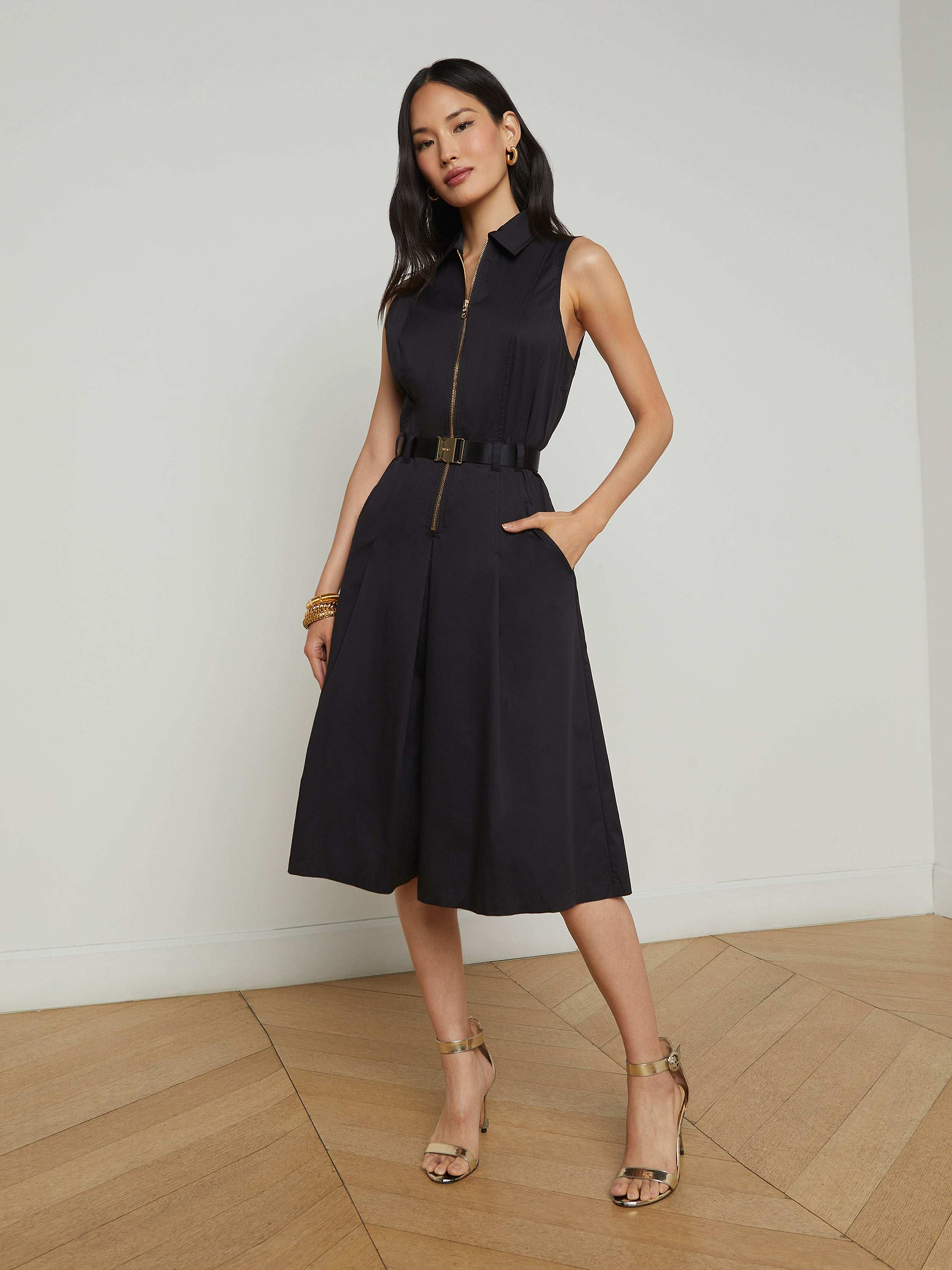 Woman wearing the Kylo Poplin Shirt Dress—a sleeveless, knee-length black dress featuring pockets and a belt—with her hands in the pockets, styled with strappy heels and gold jewelry, standing against a plain white background.