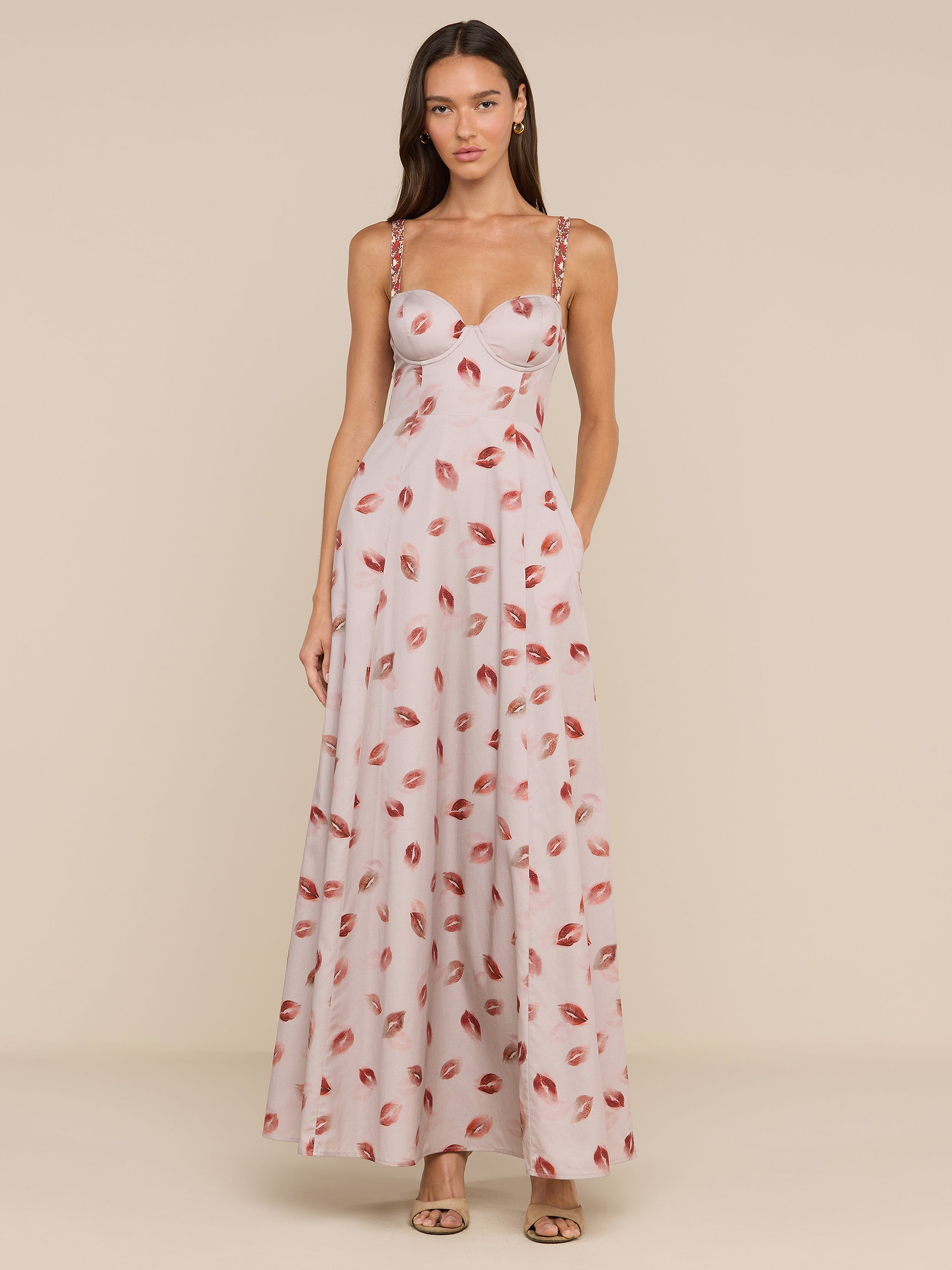 A woman stands against a plain white background wearing the Sutton Maxi Dress—a long, light pink dress featuring a lipstick-kiss print pattern and beaded straps—paired with open-toe heels.