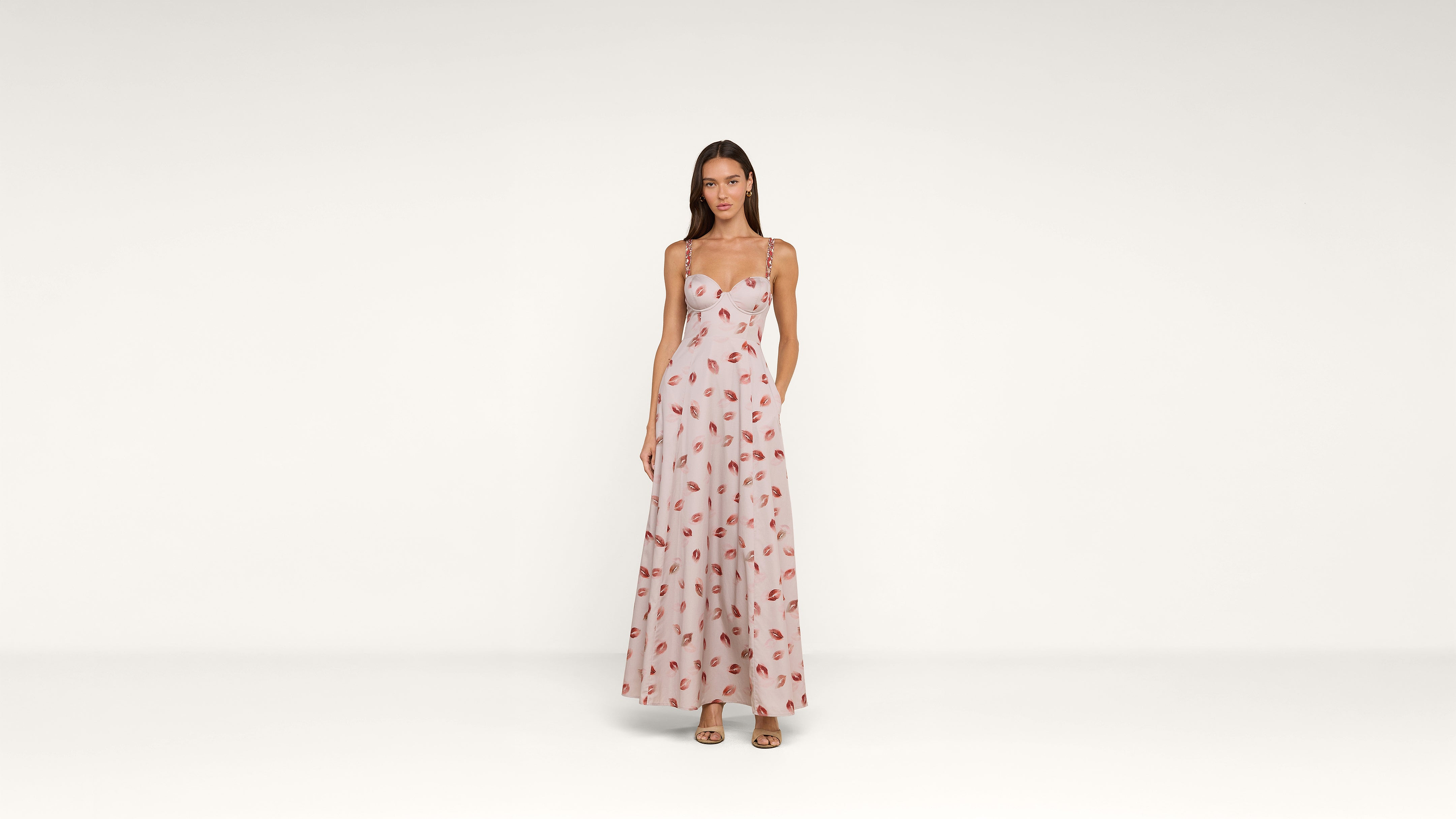 A woman stands facing forward wearing a long, light pink dress with a red lip print pattern and beige heels, against a plain white background.