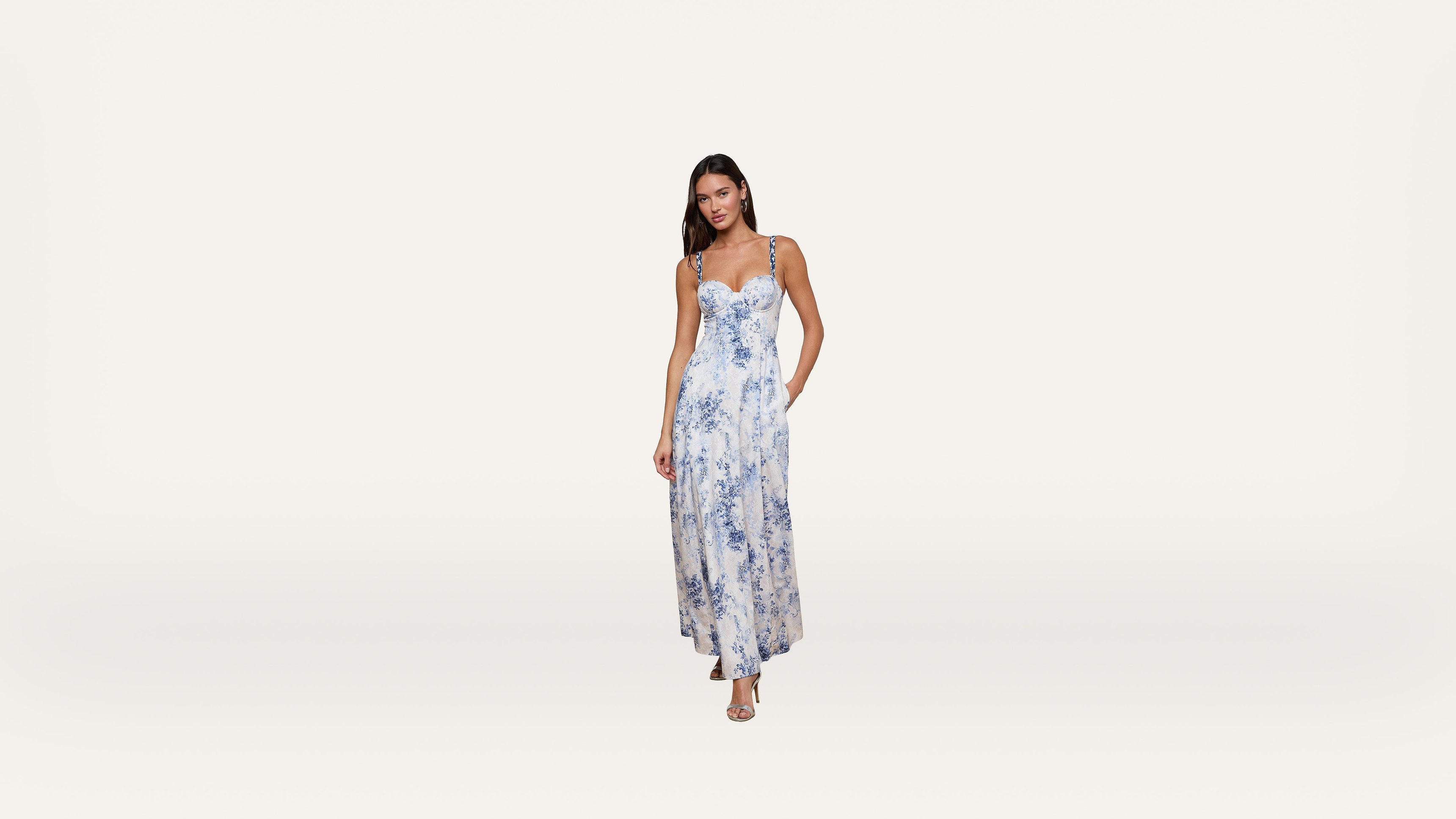 A woman stands wearing a long, sleeveless white dress with blue floral patterns and strappy sandals against a plain white background.