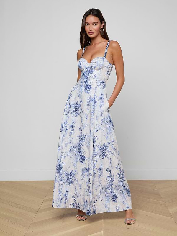 A woman wears the Sutton Maxi Dress—a long, sleeveless blue and white floral design featuring a fitted bodice, delicate beaded straps, and a flared skirt. She stands in heels against a plain white background.