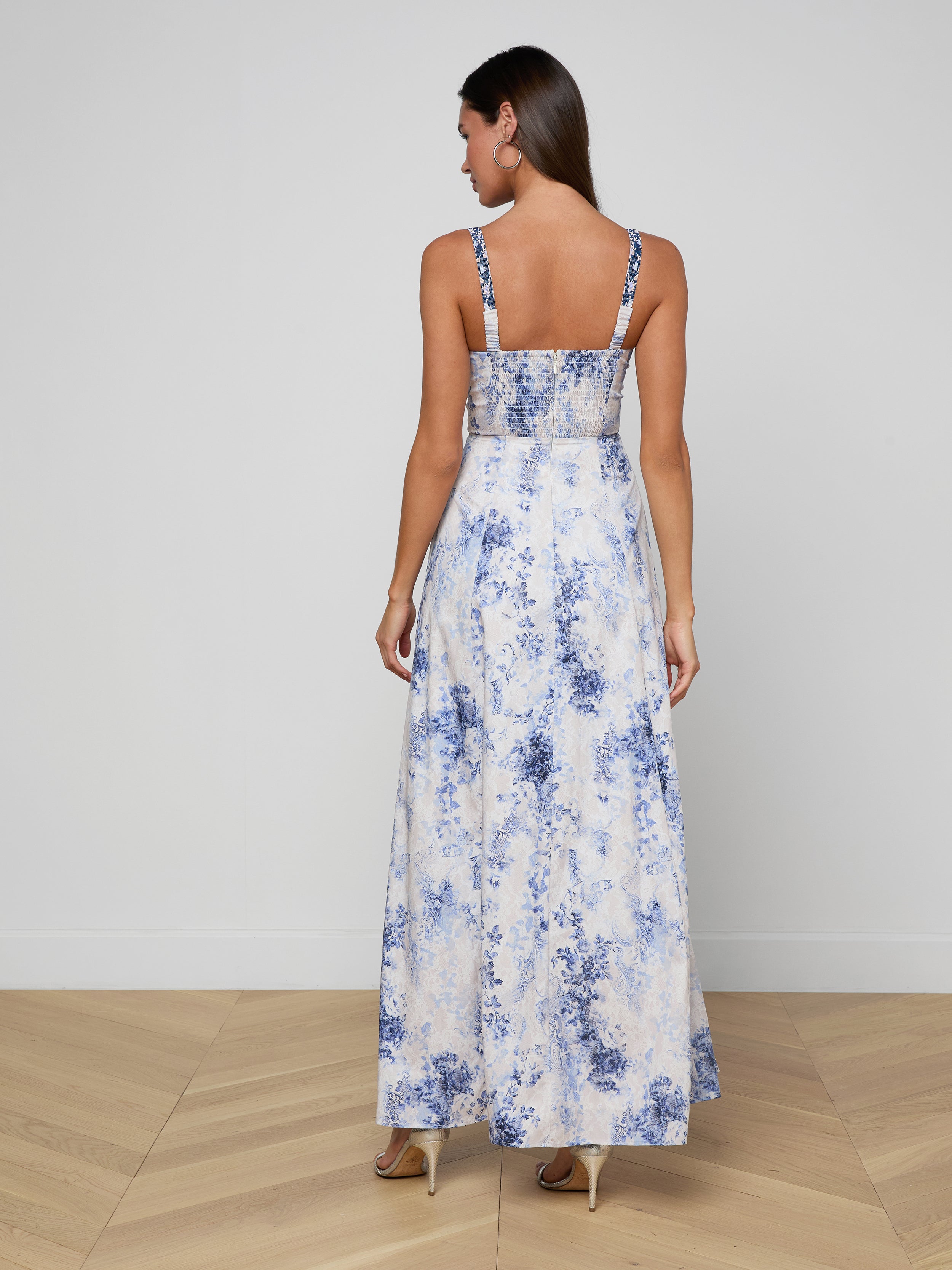 A woman stands facing away, wearing the Sutton Maxi Dress—a long, sleeveless piece with beaded straps and a blue and white pattern—paired with high heels against a plain light background.