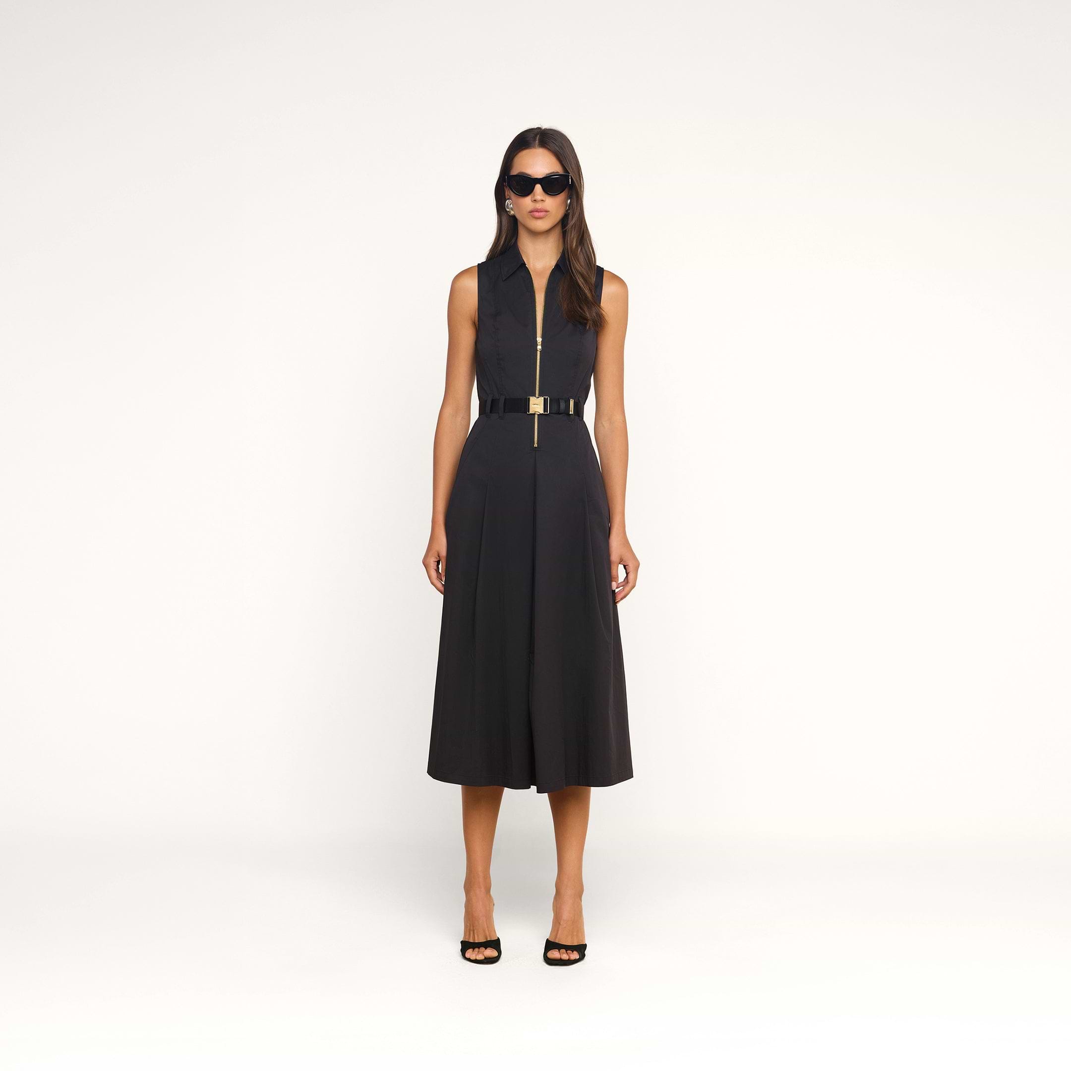 A woman wearing sunglasses and the Kolton Dress—a sleeveless black cotton-blend dress with a belt—stands against a plain white background, facing forward with her arms at her sides. She is also wearing black open-toe heels.