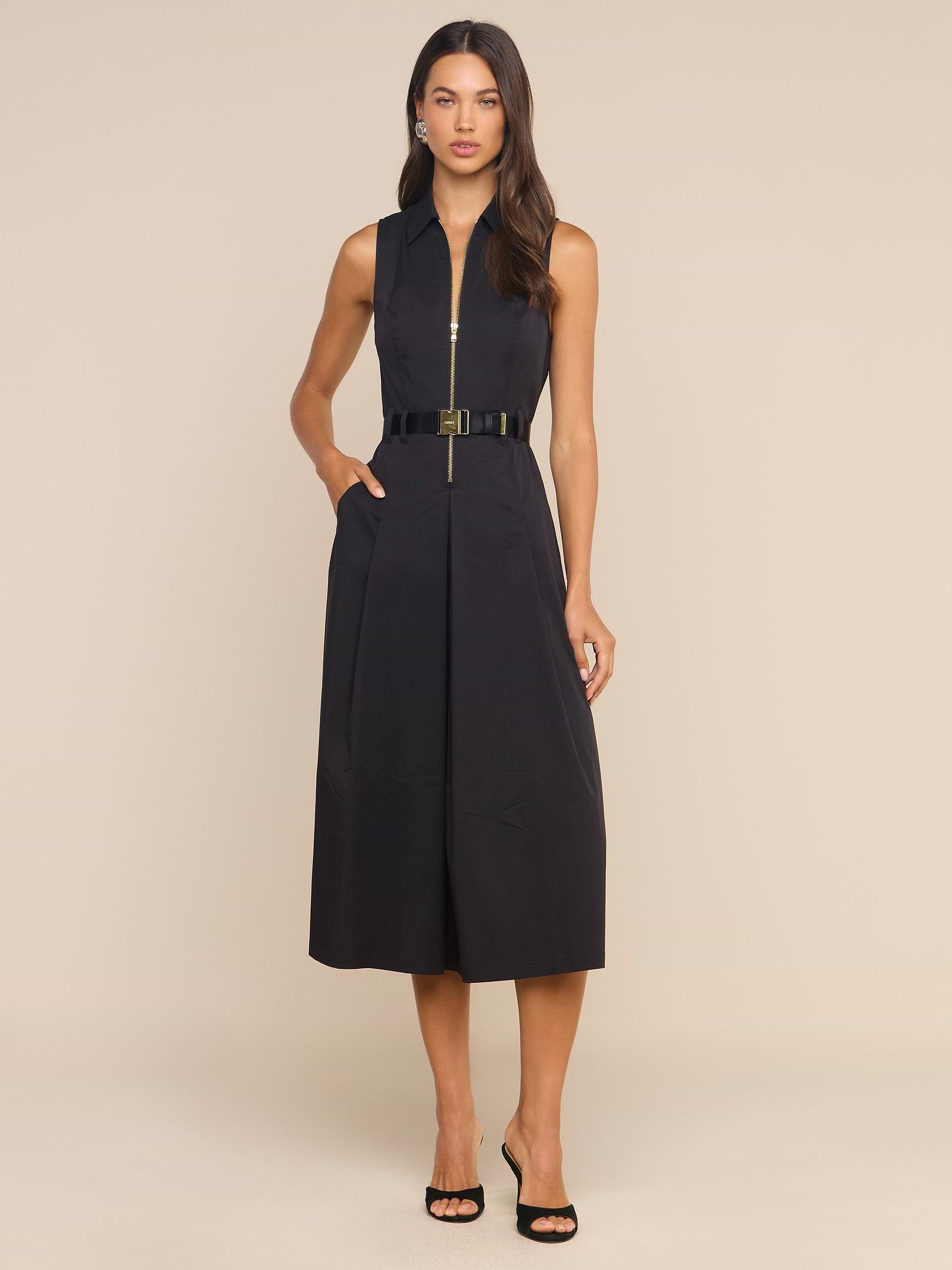 A woman stands against a plain white background wearing the Kolton Dress—a sleeveless, collared black cotton-blend midi dress with a belt—paired with black heeled sandals.