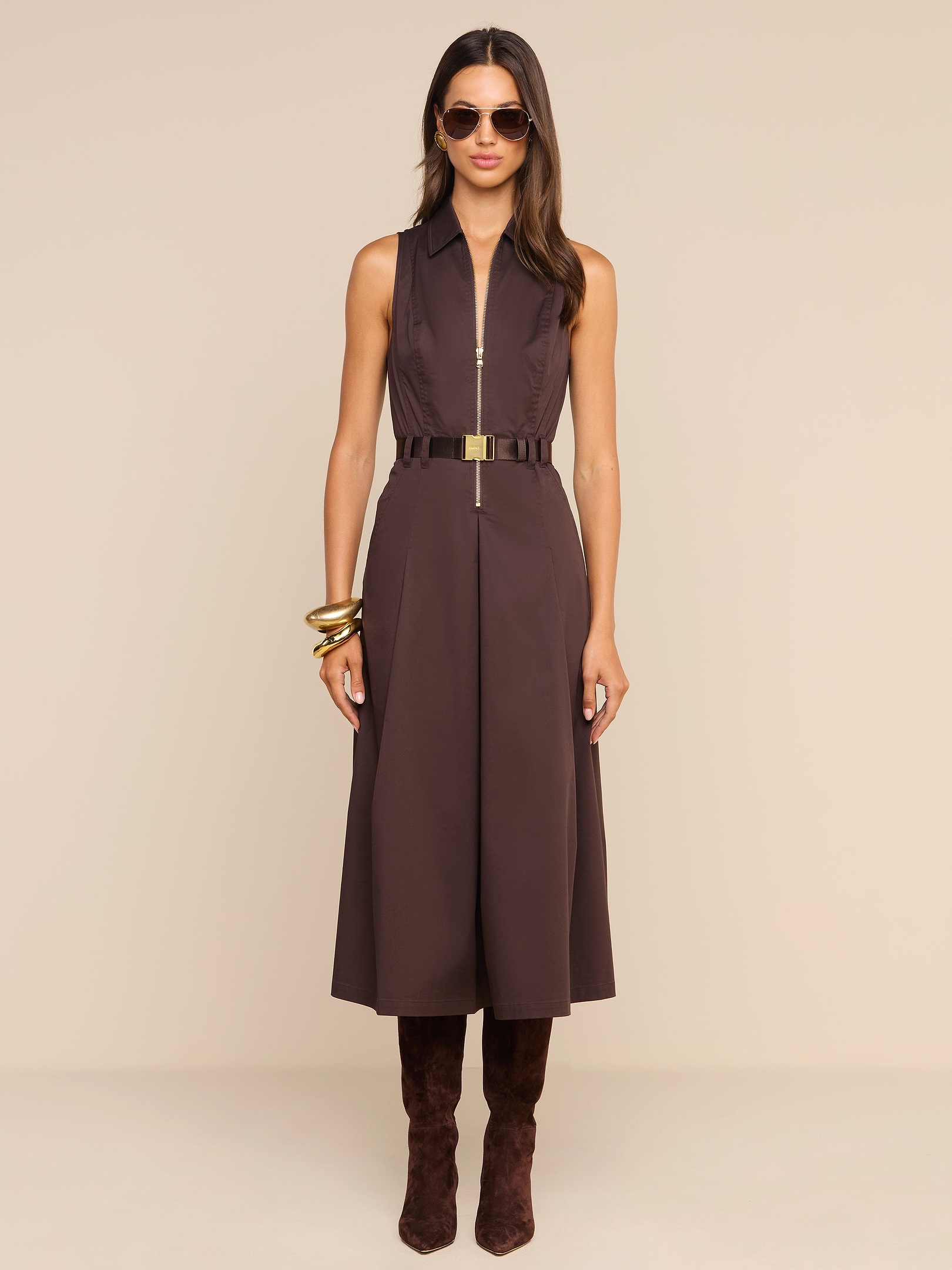 A woman wearing the Kolton Dress—a sleeveless brown cotton-blend poplin midi dress—styled with a gold belt, large bangles, sunglasses, and knee-high brown boots stands against a plain white background.
