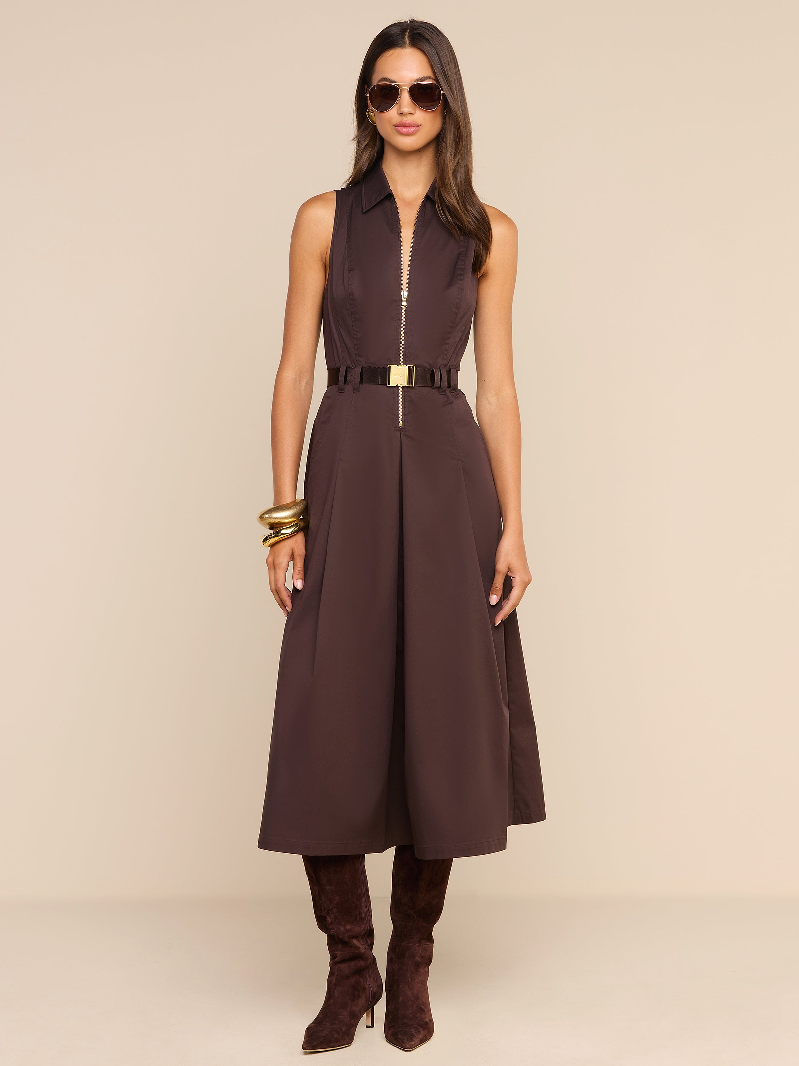 Woman wearing the Kolton Dress—a sleeveless brown cotton-blend poplin midi dress—styled with a gold belt, large gold bangles, dark brown boots, and sunglasses, standing against a plain white background.