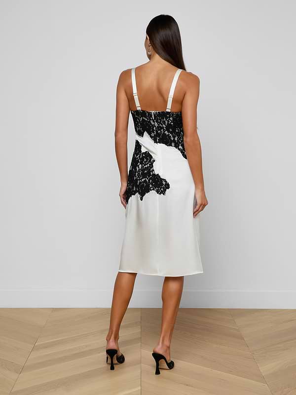 A woman stands facing away, wearing the knee-length Fenna Silk Lace Dress with a bustier top and black high heels, against a plain light background.