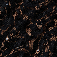 Close-up of the Lindsay Sequined Lace Dress, showcasing its intricate black lace fabric with a floral pattern, accented by small sequins and a nude-colored backing visible through the open mesh—perfect for an elegant off-shoulder evening look.