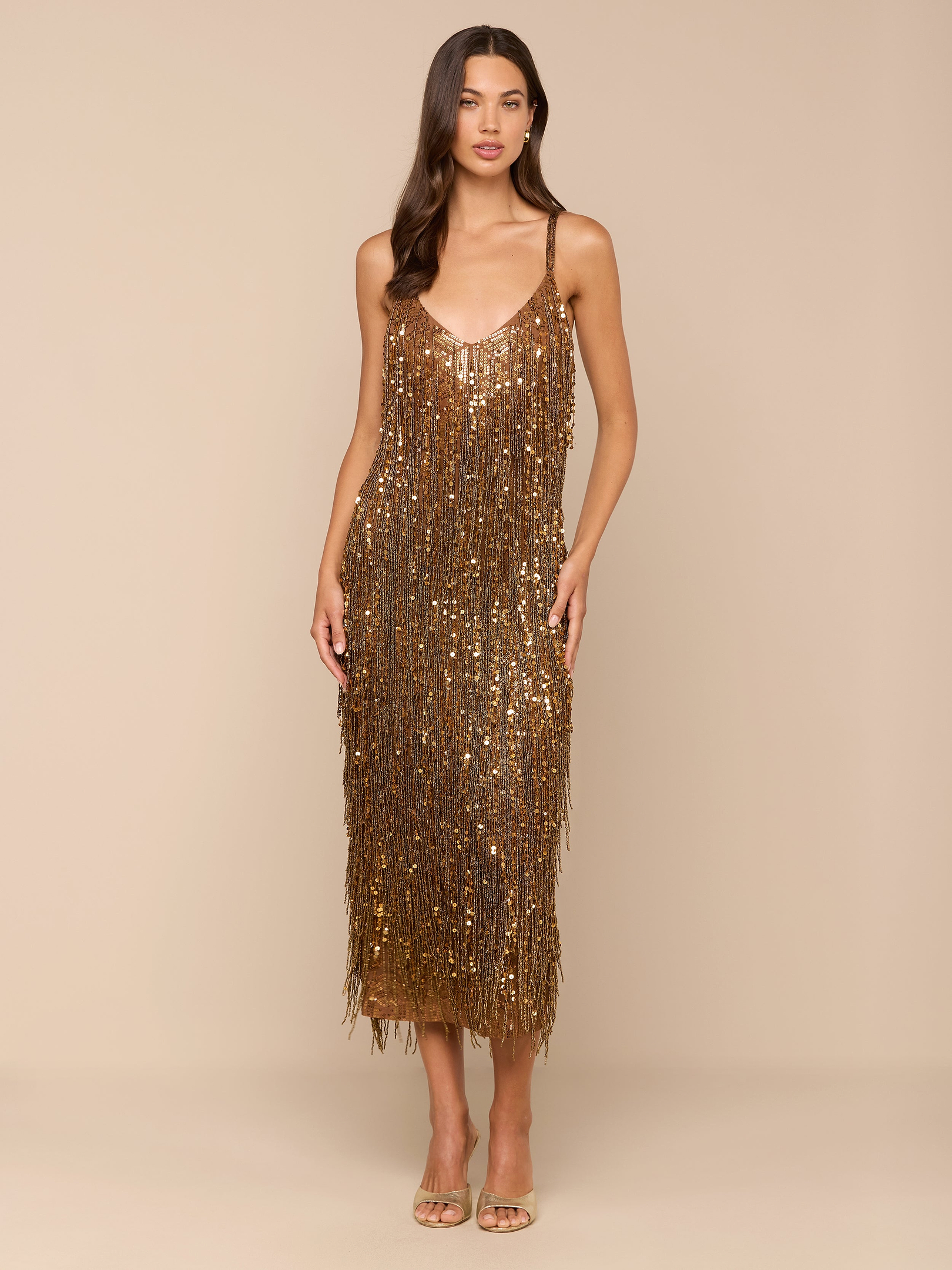 Une femme aux longs cheveux bruns se tient debout sur un fond beige, portant la robe à franges à paillettes Noella - dorée avec des détails de franges et de fines bretelles - et des talons métalliques. Elle a une expression neutre et ses bras sont détendus le long du corps.