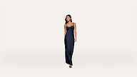 A woman in a long, dark navy satin dress with spaghetti straps stands against a plain white background, looking slightly to her left.