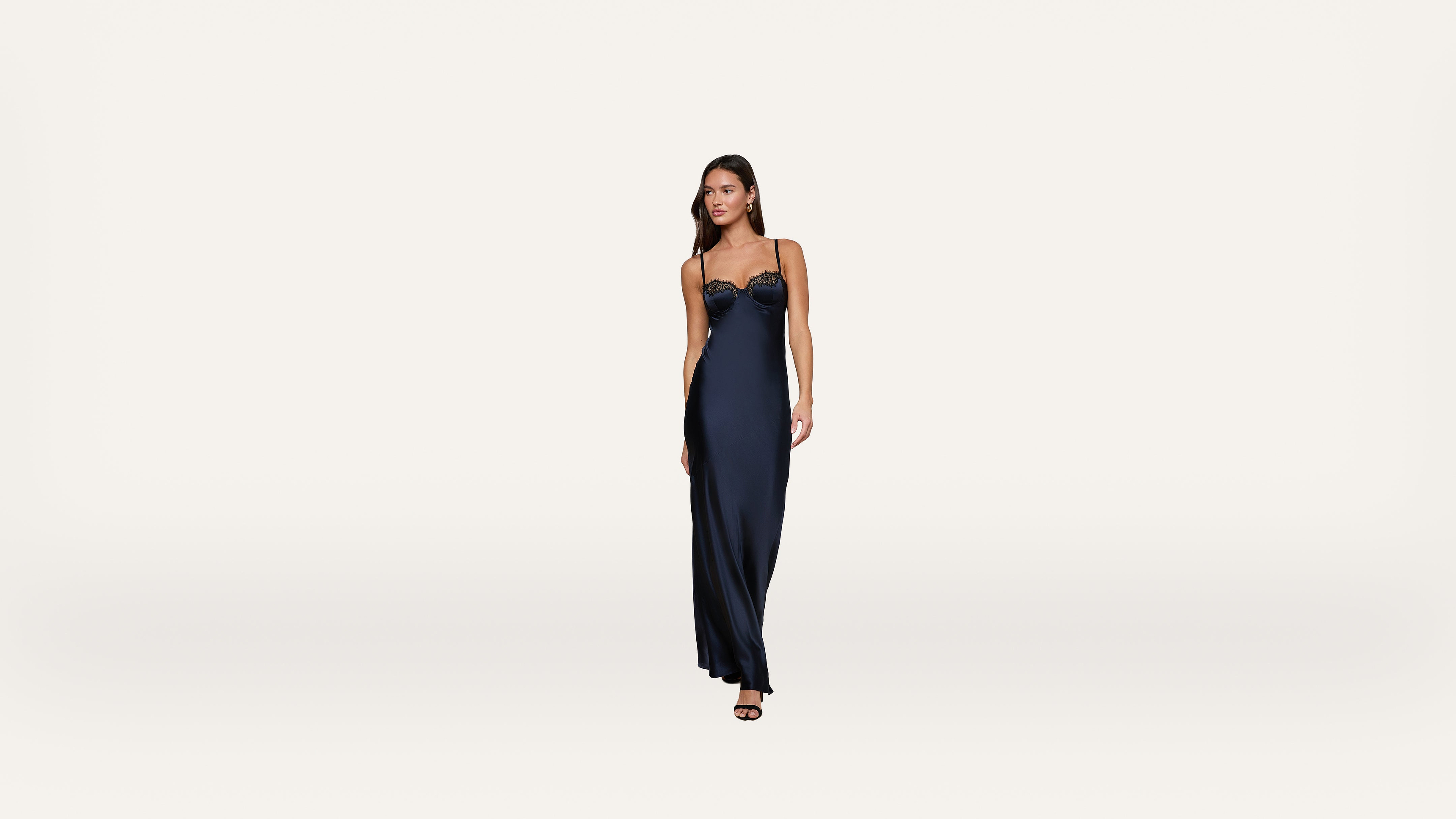 A woman in a long, dark navy satin dress with spaghetti straps stands against a plain white background, looking slightly to her left.