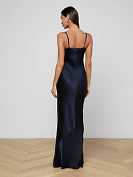 A woman stands facing away from the camera against a plain white background, wearing the Shayla Silk Lace Gown—a long, dark blue dress with thin straps crafted from satin and delicate lace.