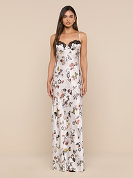 A woman stands facing forward against a plain white background, wearing the Sayla Silk Gown—a long, white silk dress featuring a floral and butterfly lock print with black lace trim along the neckline.