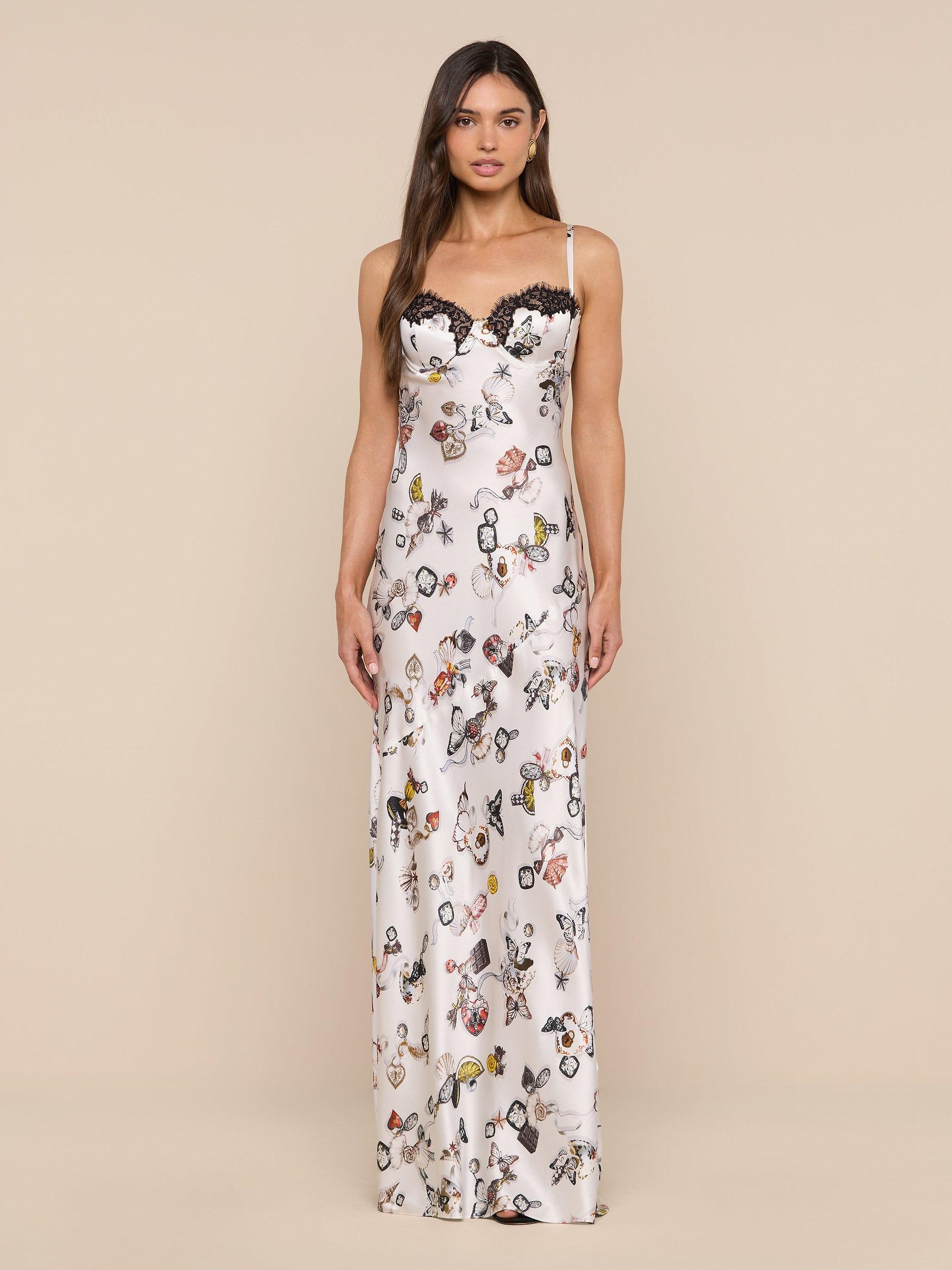 A woman stands facing forward against a plain white background, wearing the Sayla Silk Gown—a long, white silk dress featuring a floral and butterfly lock print with black lace trim along the neckline.