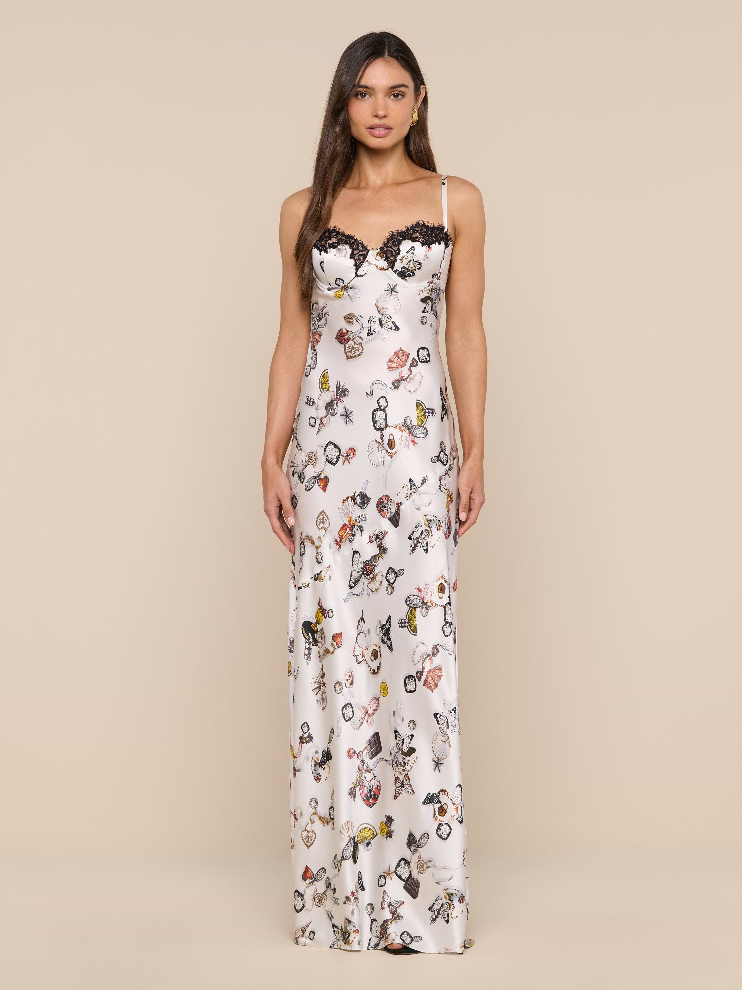 A woman stands facing forward against a plain white background, wearing the Sayla Silk Gown—a long, white silk dress featuring a floral and butterfly lock print with black lace trim along the neckline.