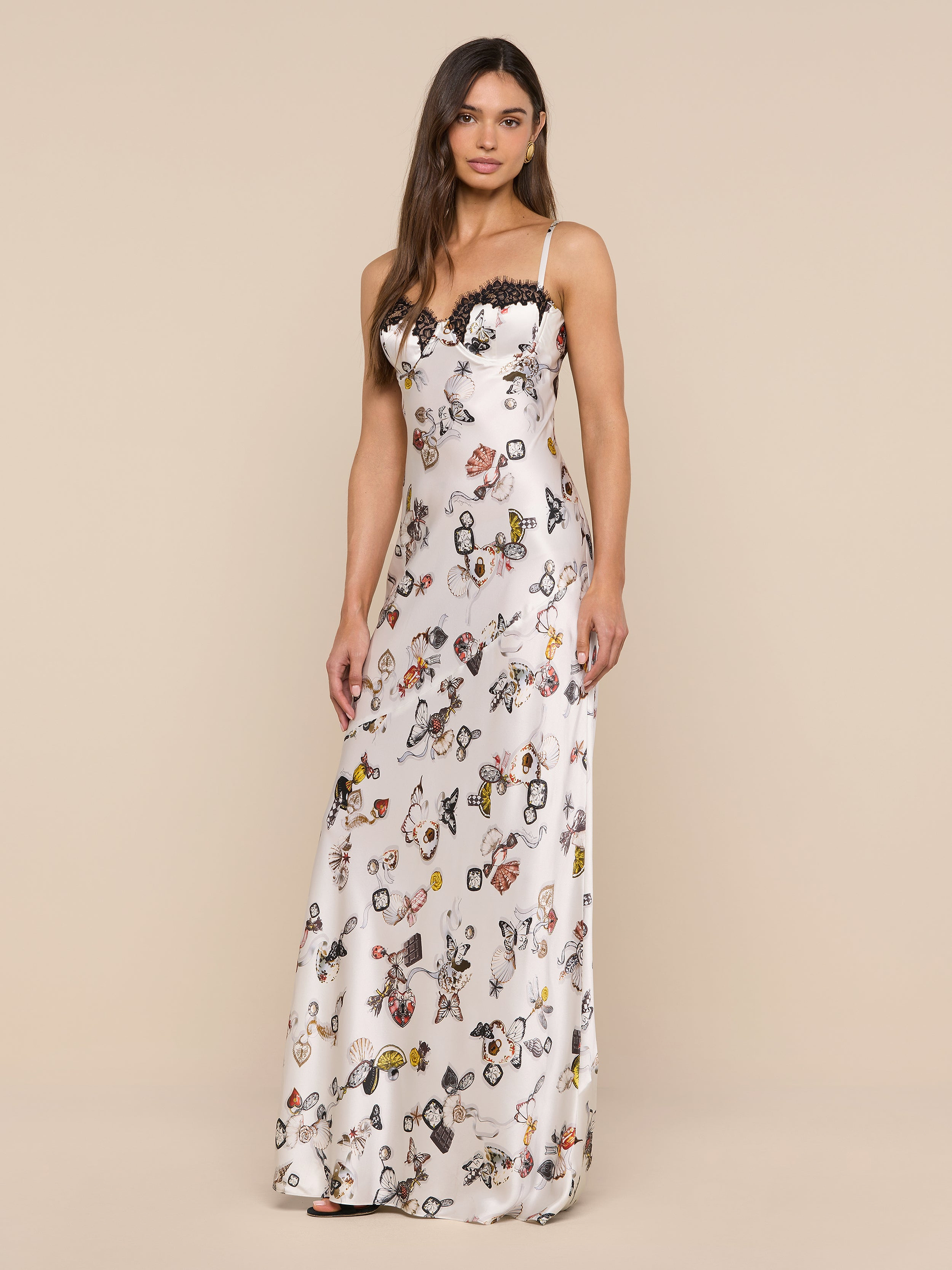 A woman stands against a plain white background, wearing the Sayla Silk Gown—an elegant, long, sleeveless dress featuring a butterfly lock print and delicate lace trim.