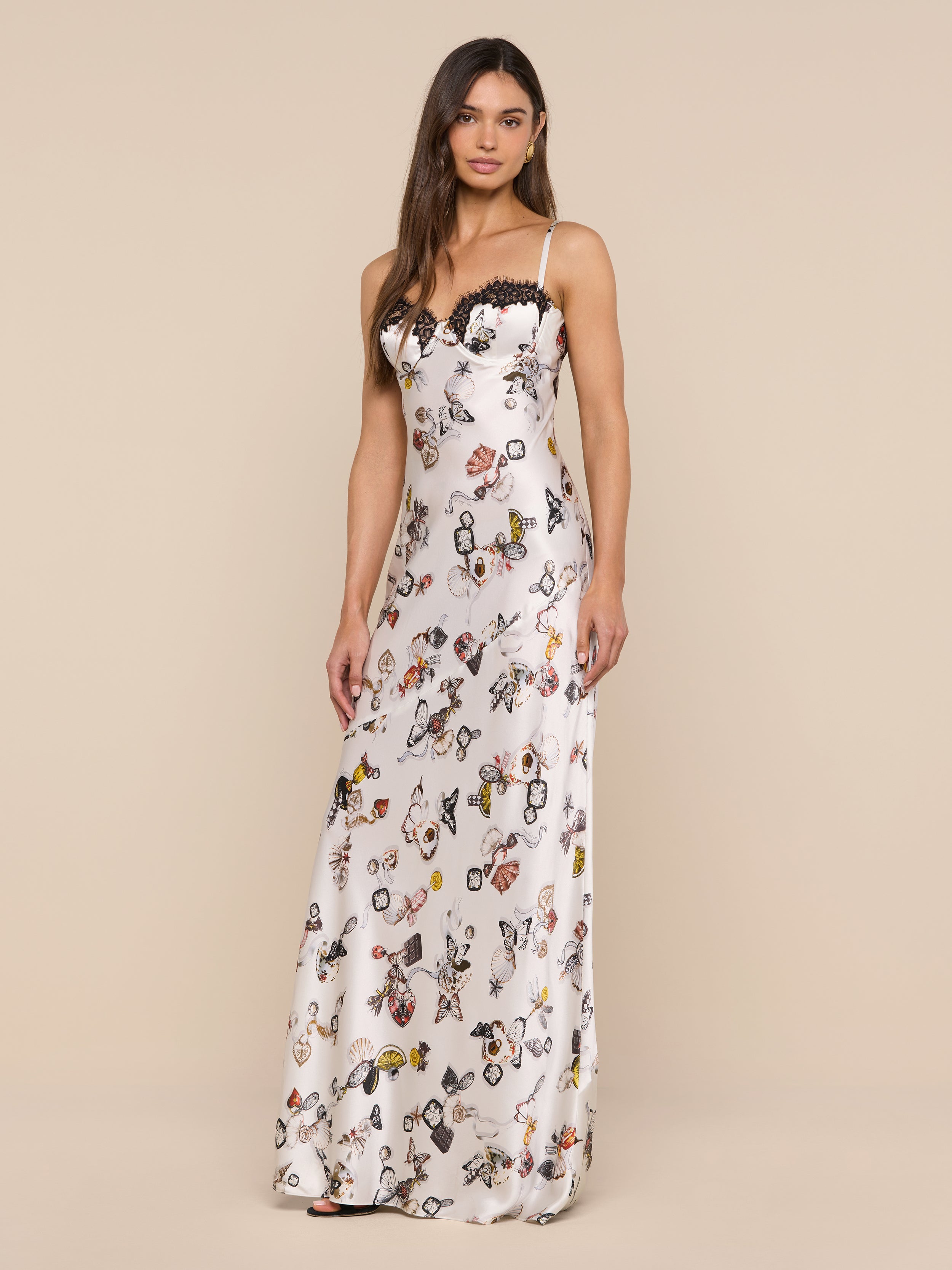 A woman stands against a plain white background, wearing the Sayla Silk Gown—an elegant, long, sleeveless dress featuring a butterfly lock print and delicate lace trim.