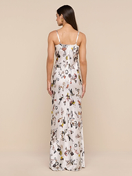 A woman stands against a plain background, facing away, wearing the Sayla Silk Gown—a long white silk dress with thin straps and vibrant illustrated patterns that evoke vintage glamour. Her straight hair falls down her back.