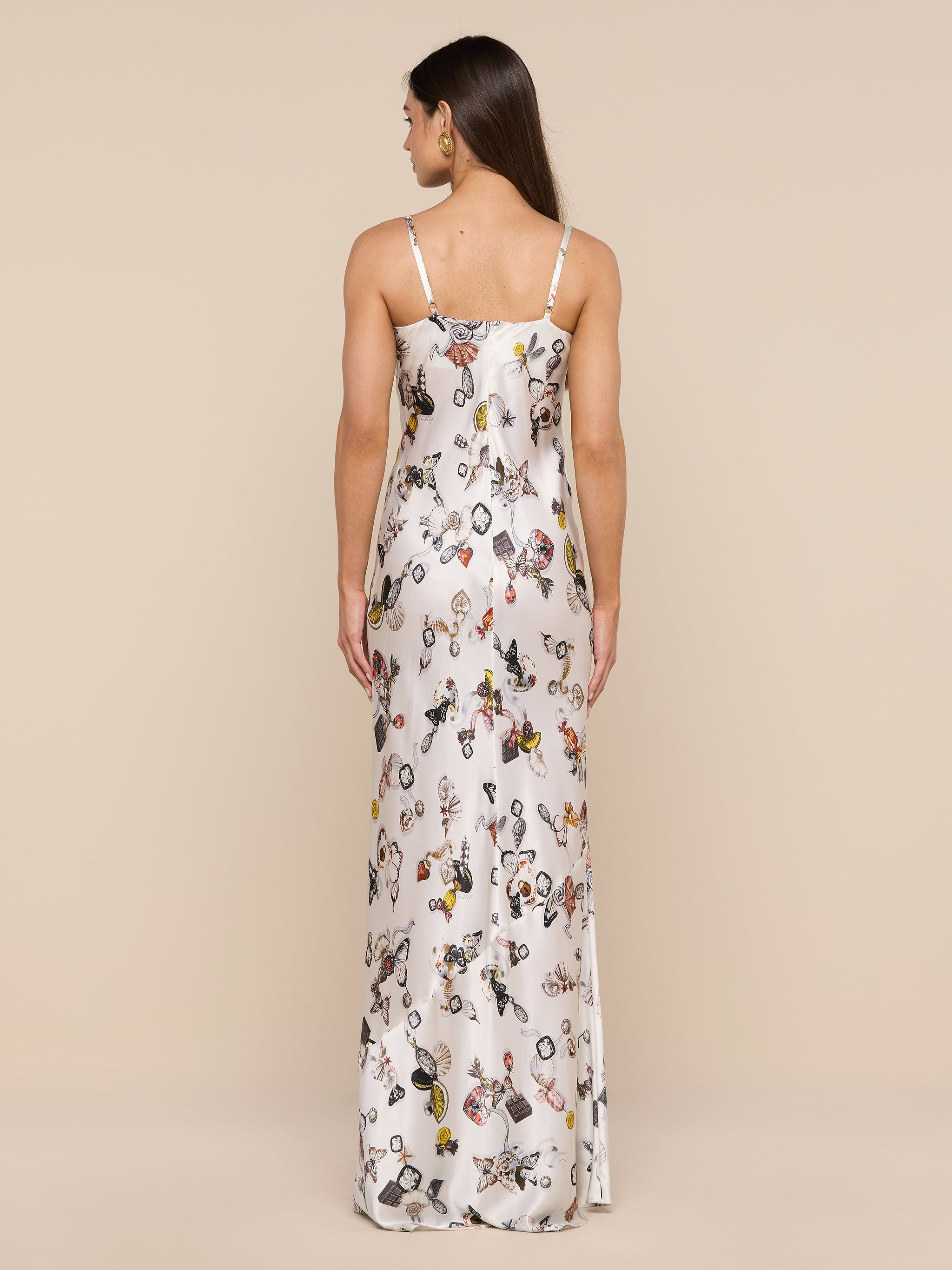 A woman stands against a plain background, facing away, wearing the Sayla Silk Gown—a long white silk dress with thin straps and vibrant illustrated patterns that evoke vintage glamour. Her straight hair falls down her back.
