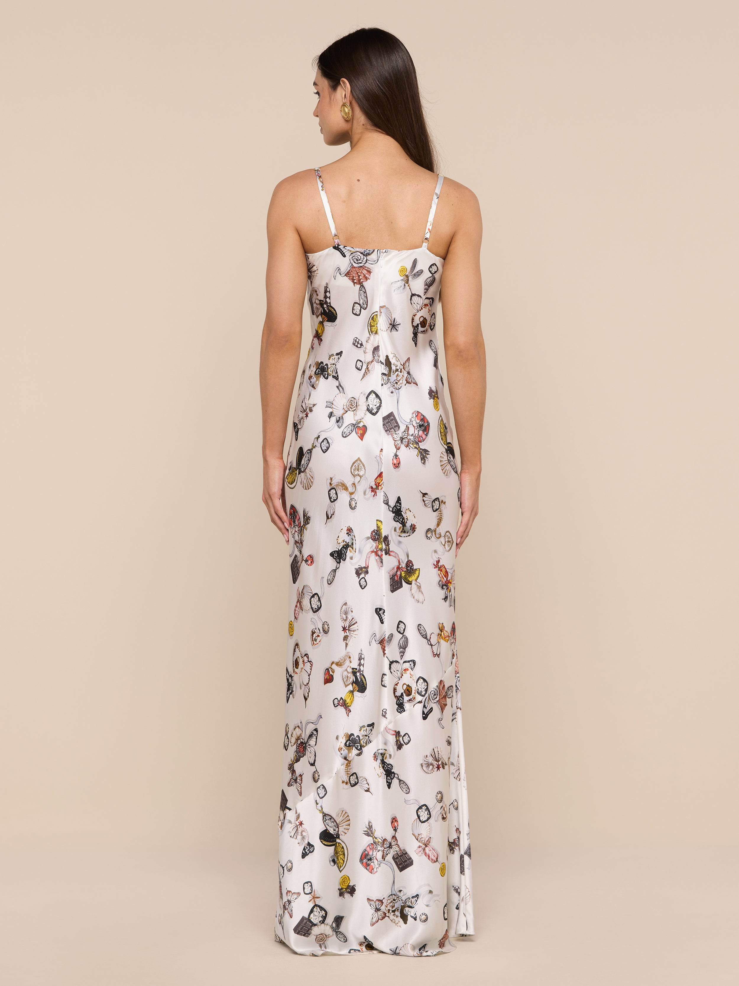 A woman stands against a plain background, facing away, wearing the Sayla Silk Gown—a long white silk dress with thin straps and vibrant illustrated patterns that evoke vintage glamour. Her straight hair falls down her back.