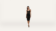 A woman wearing a black lace dress and black heels stands against a plain white background, posing and looking toward the camera.