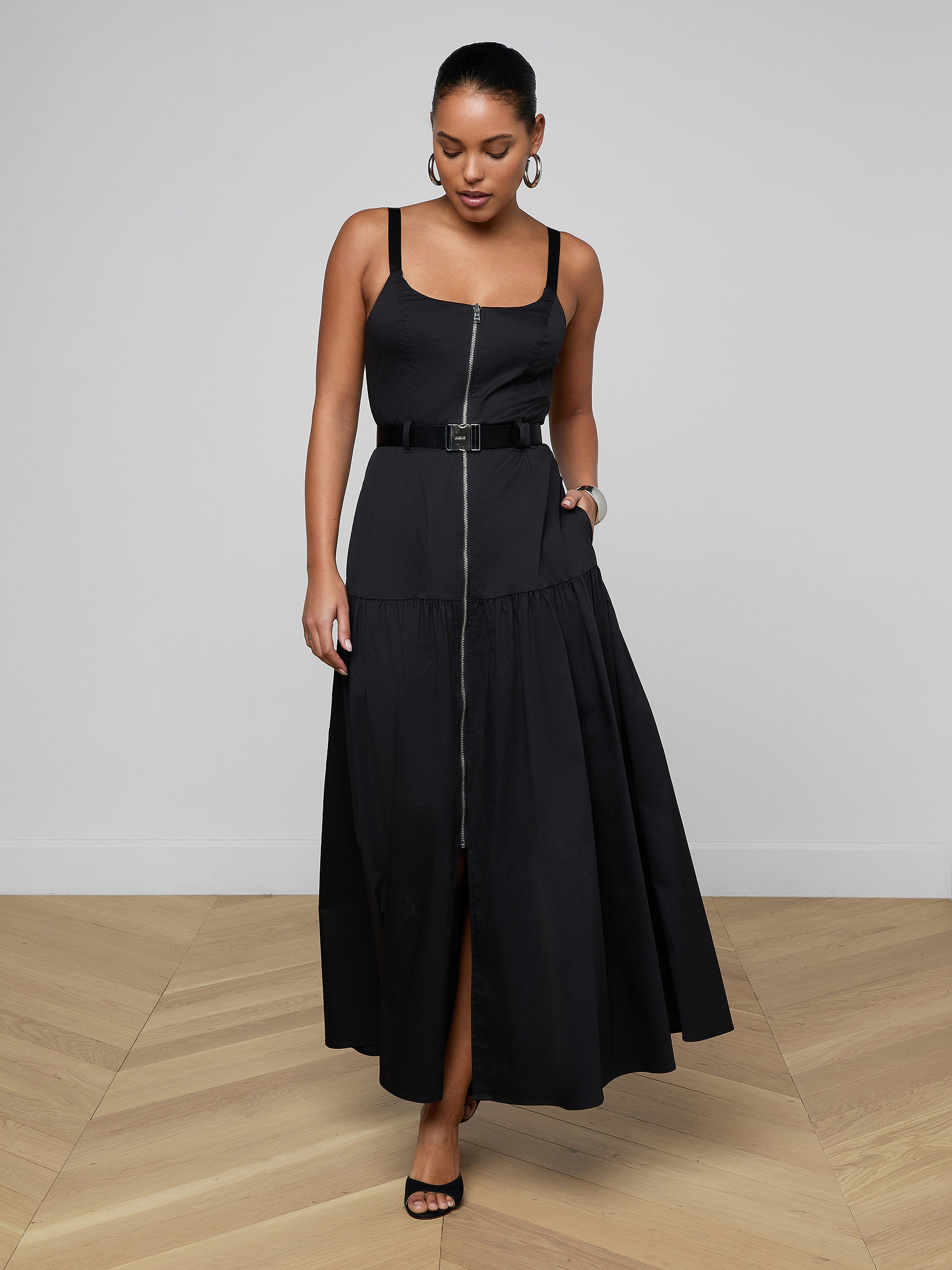 A woman wearing the Rima Maxi Dress—a sleeveless black maxi dress with a belted waist crafted from stretch poplin—paired with black heels stands against a plain light background, looking down with one hand touching the skirt.