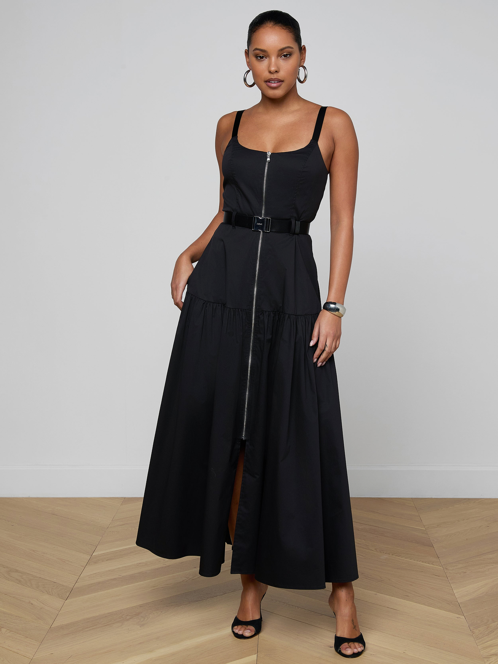 A woman stands against a plain light background wearing the Rima Maxi Dress—a sleeveless black stretch poplin maxi dress with a square neckline—paired with open-toe heels, hoop earrings, and a bracelet.