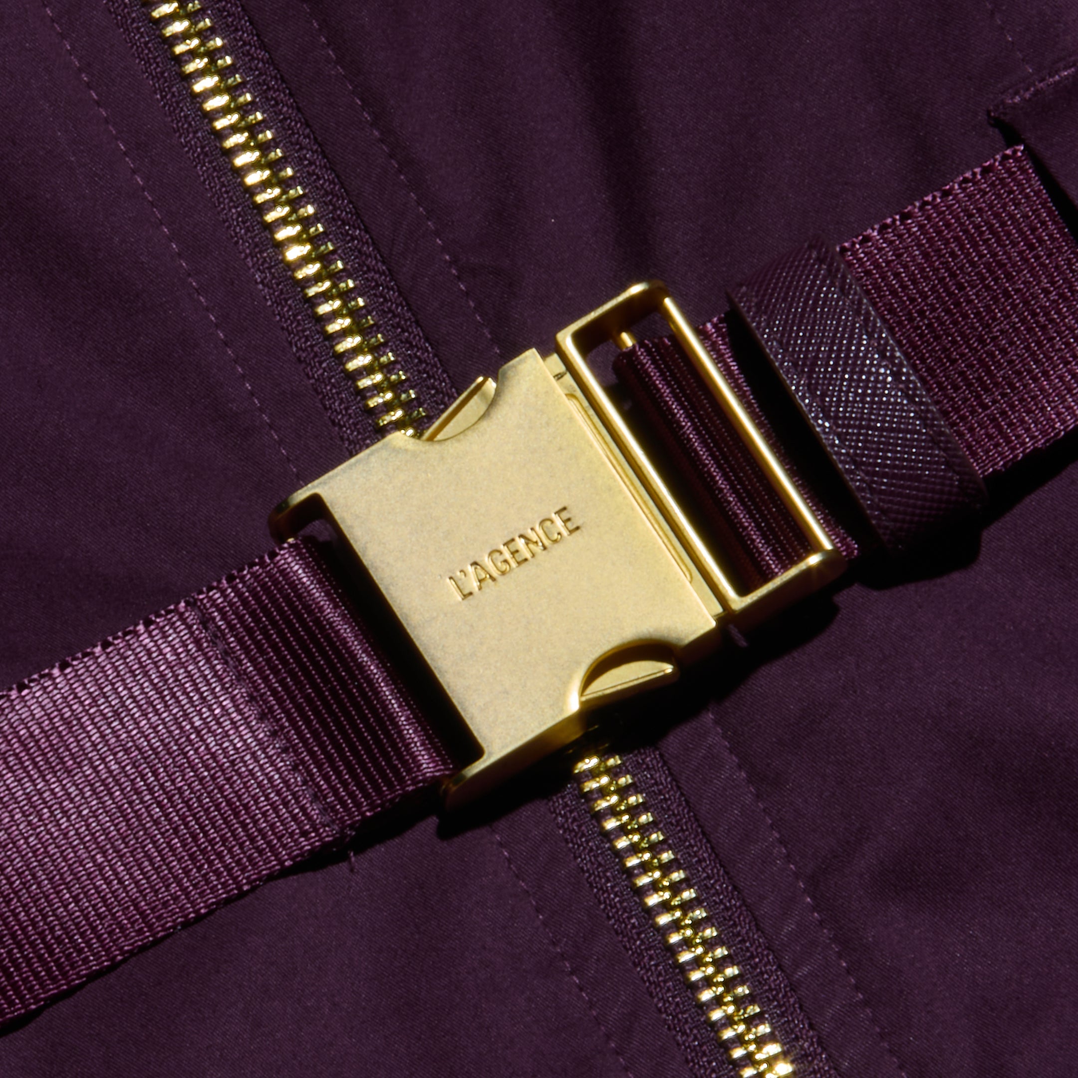 An adjustable belt made from a dark purple strap, featuring a gold metal buckle engraved with LAGENCE, accents the Rima Dress, shown on matching fabric beside a gold zipper.