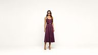 A woman wearing a sleeveless purple dress, sunglasses, and heels stands against a plain white background, facing forward with her arms relaxed by her sides.